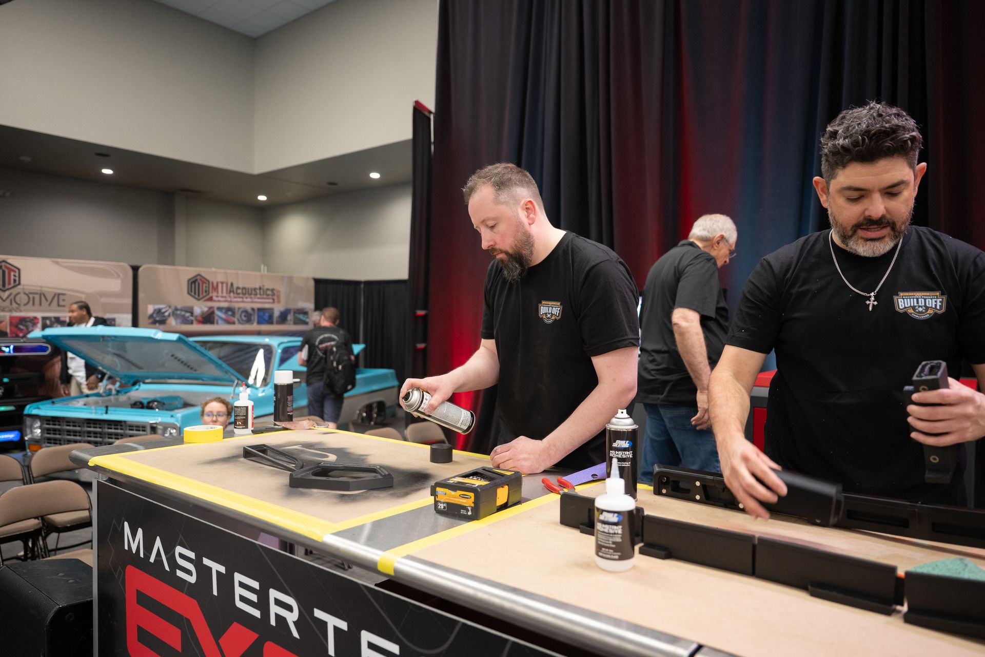 Men working on car parts at a trade show booth, one spraying paint.
