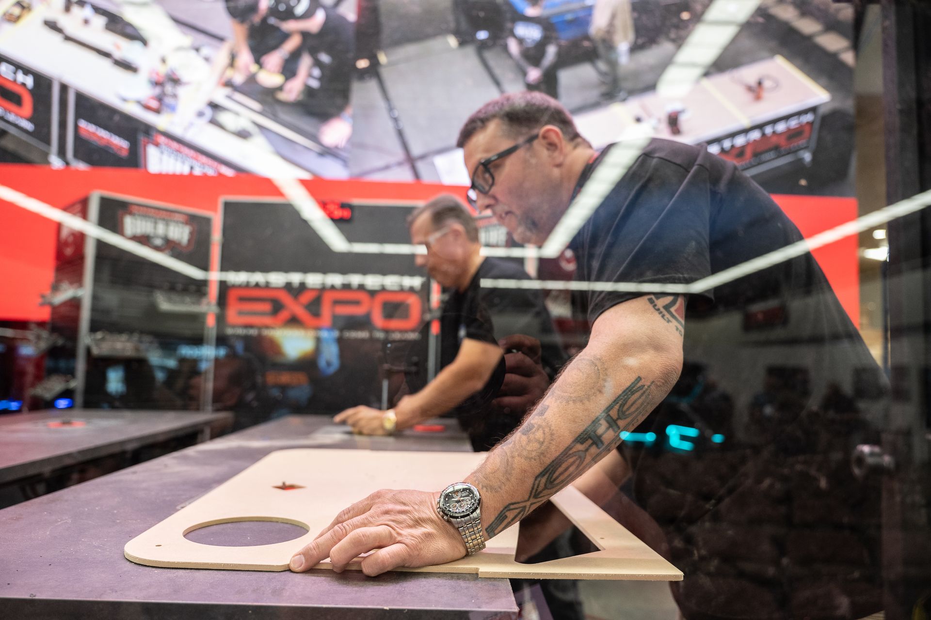 Two men working at a table, hands on a wooden sheet. Fair in the background.