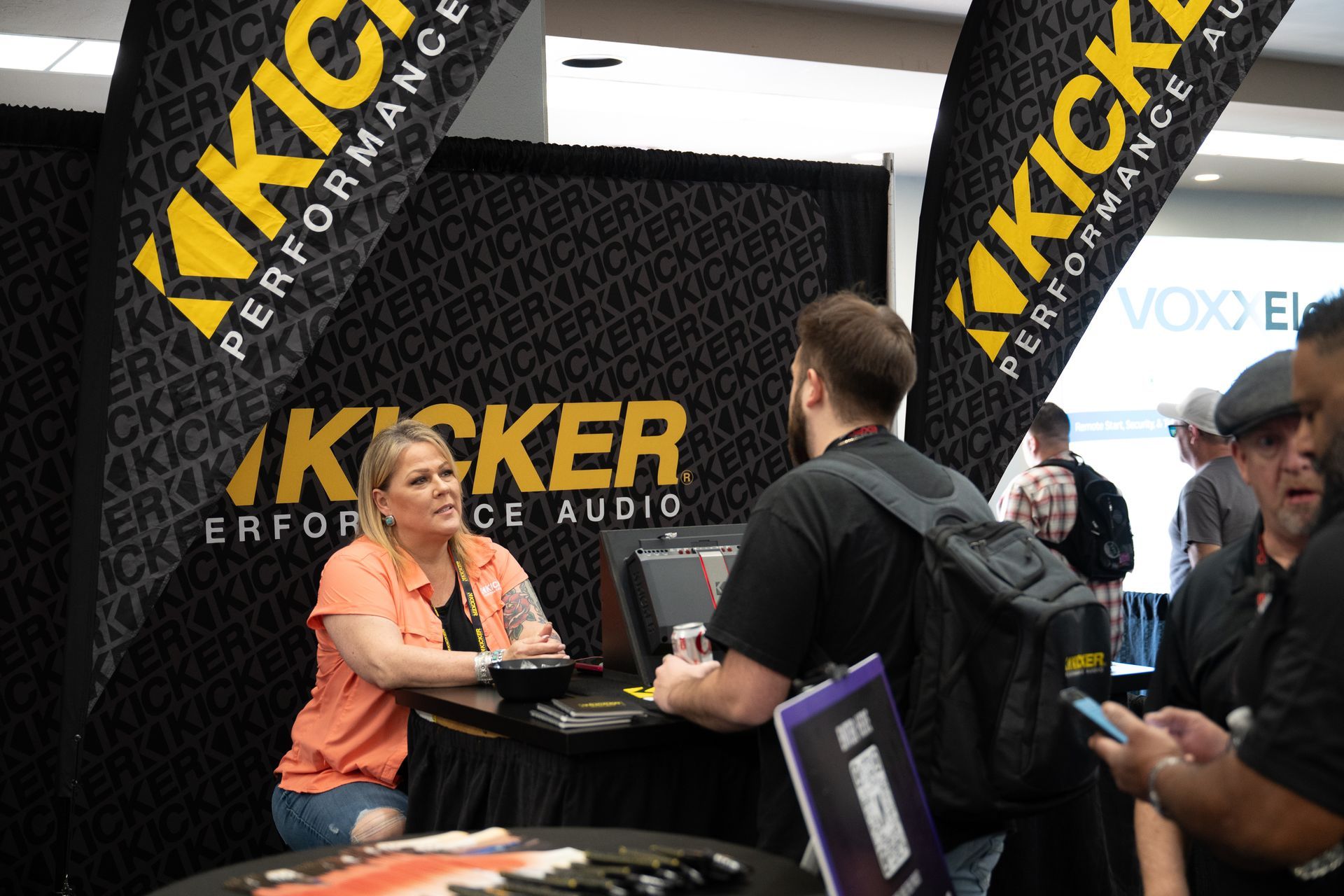 People at a Kicker Performance Audio booth; a woman and a man interact at a table, banners overhead.