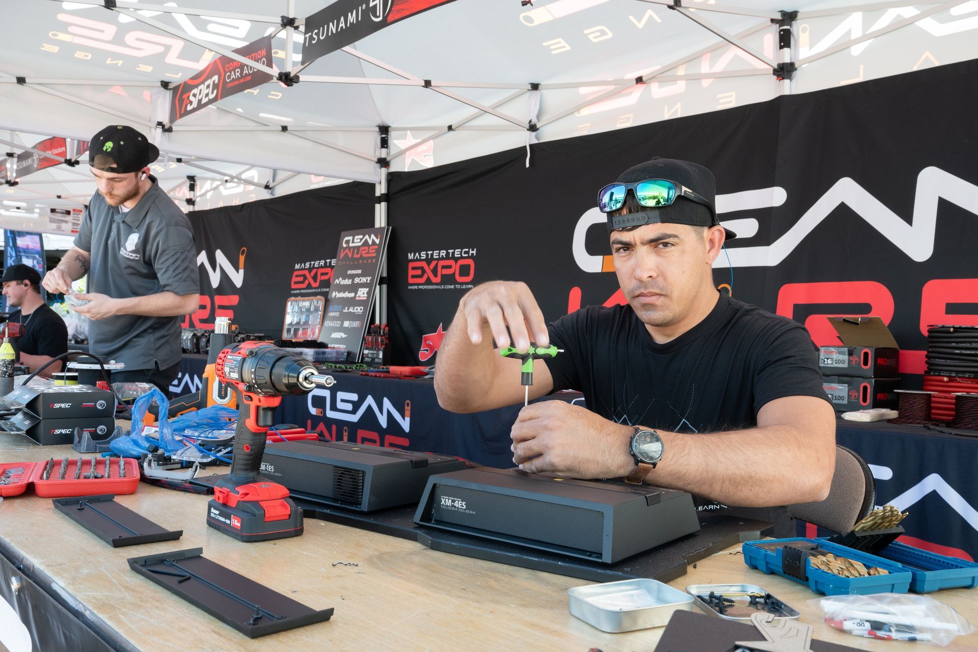 Two men working at a booth, one using a screwdriver, the other assembling parts, Clean Garage Expo.