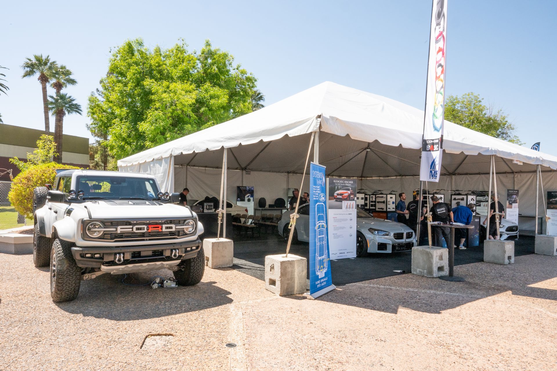 White Ford Bronco parked outside a white tent at an outdoor event. People stand inside.