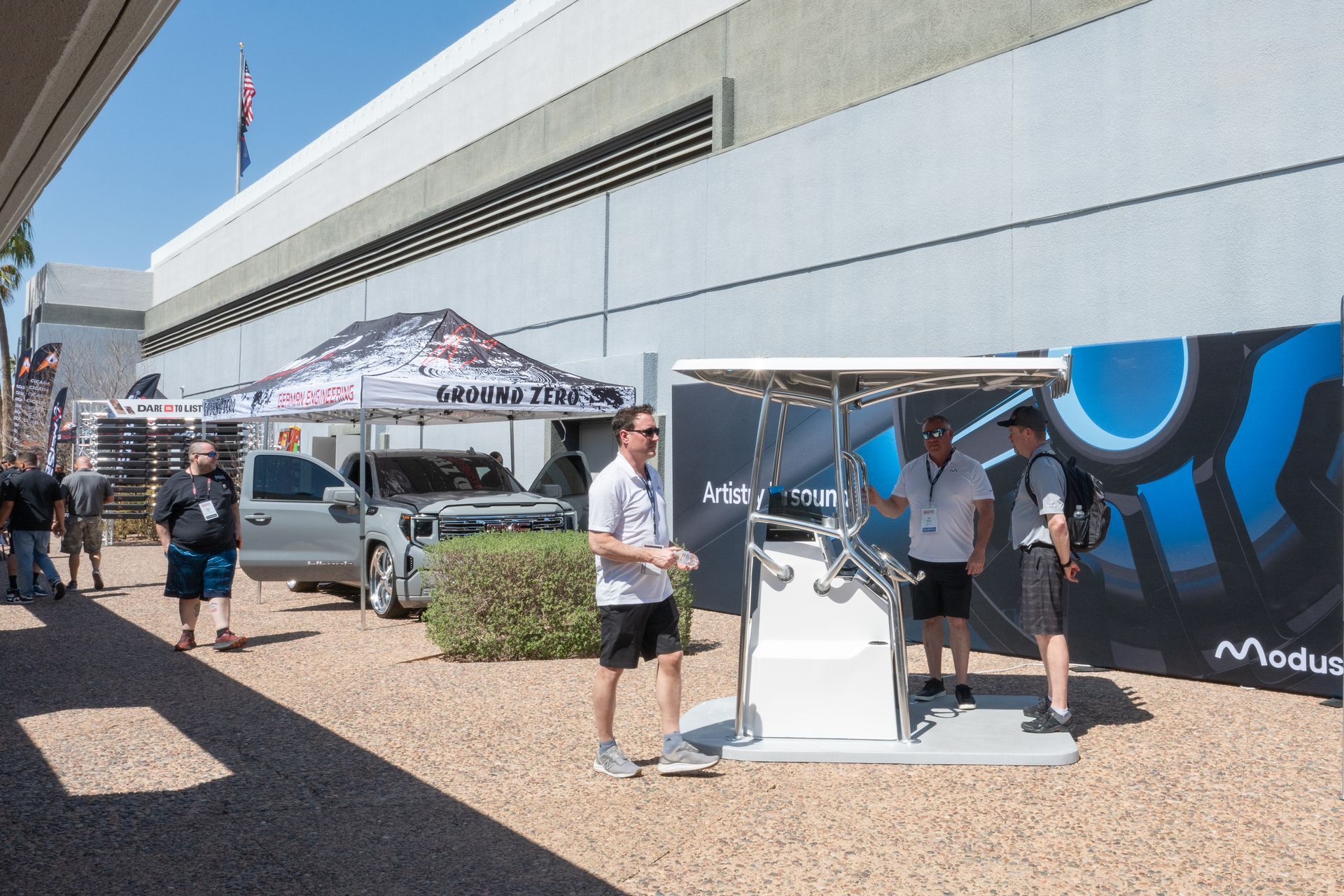 People stand near a white vehicle display with a promotional tent on a sunny day.