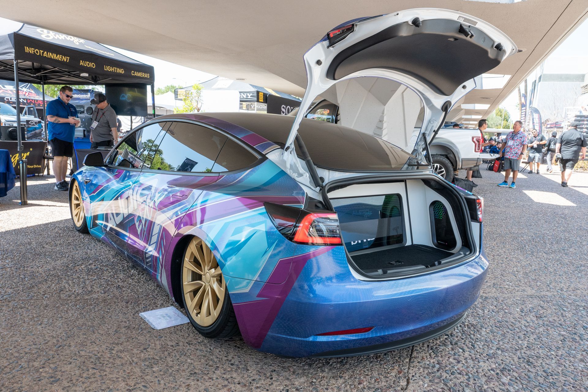 Customized blue Tesla with gold wheels, trunk open at an outdoor event.