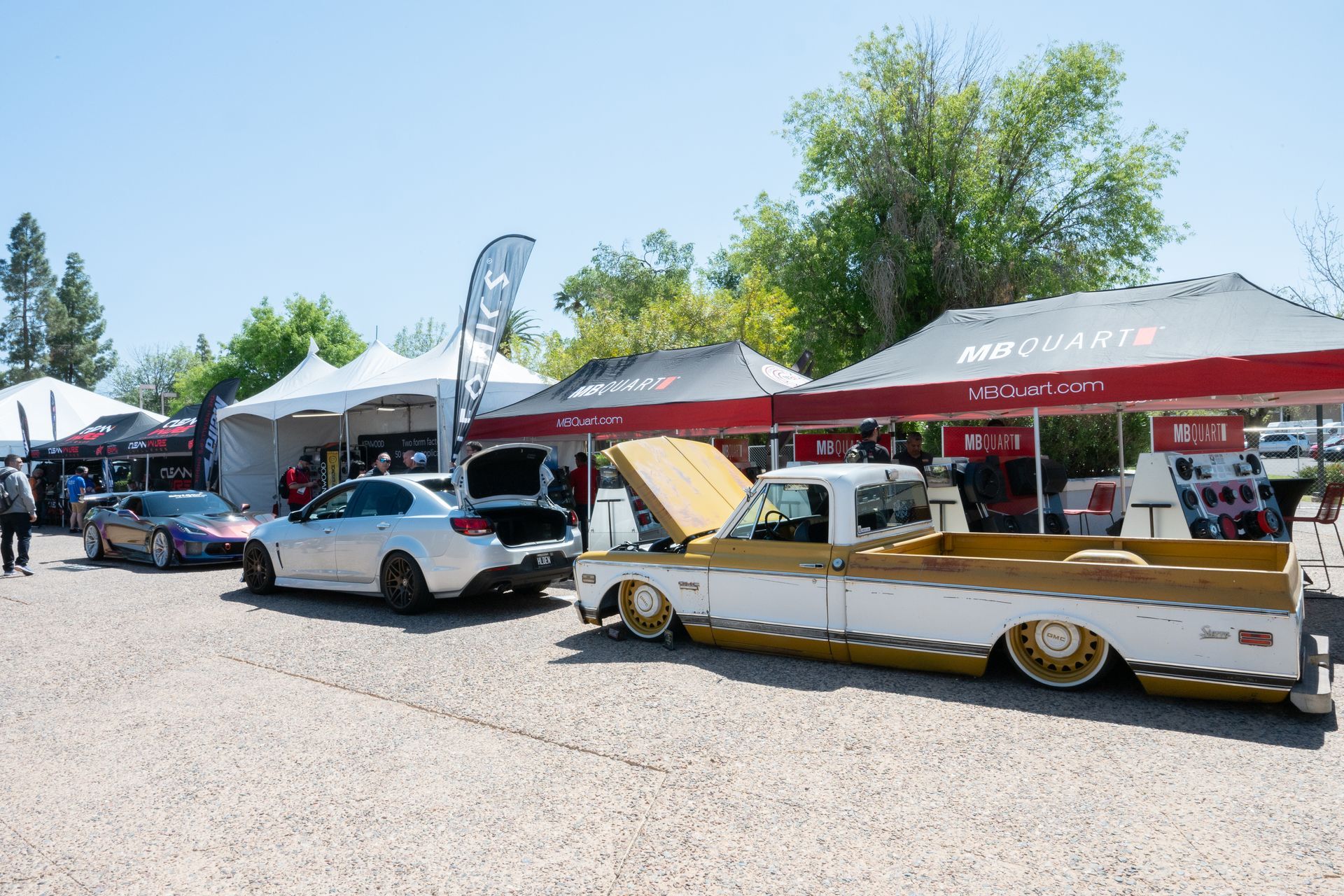 Car show with various vehicles on display next to vendor tents on a sunny day.