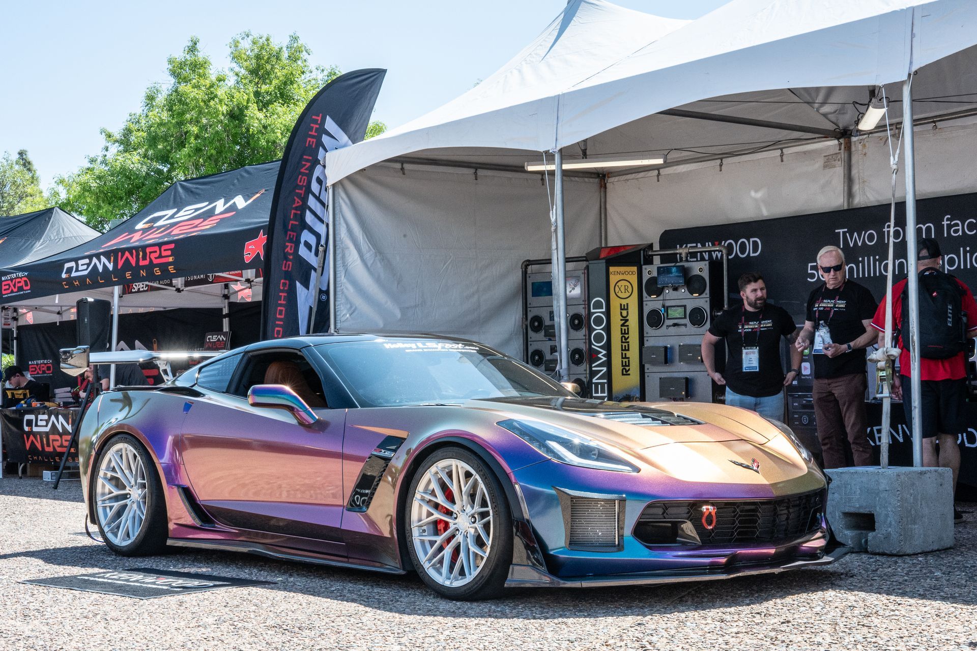Custom iridescent Corvette sports car at an outdoor event, with two people in background near a tent.
