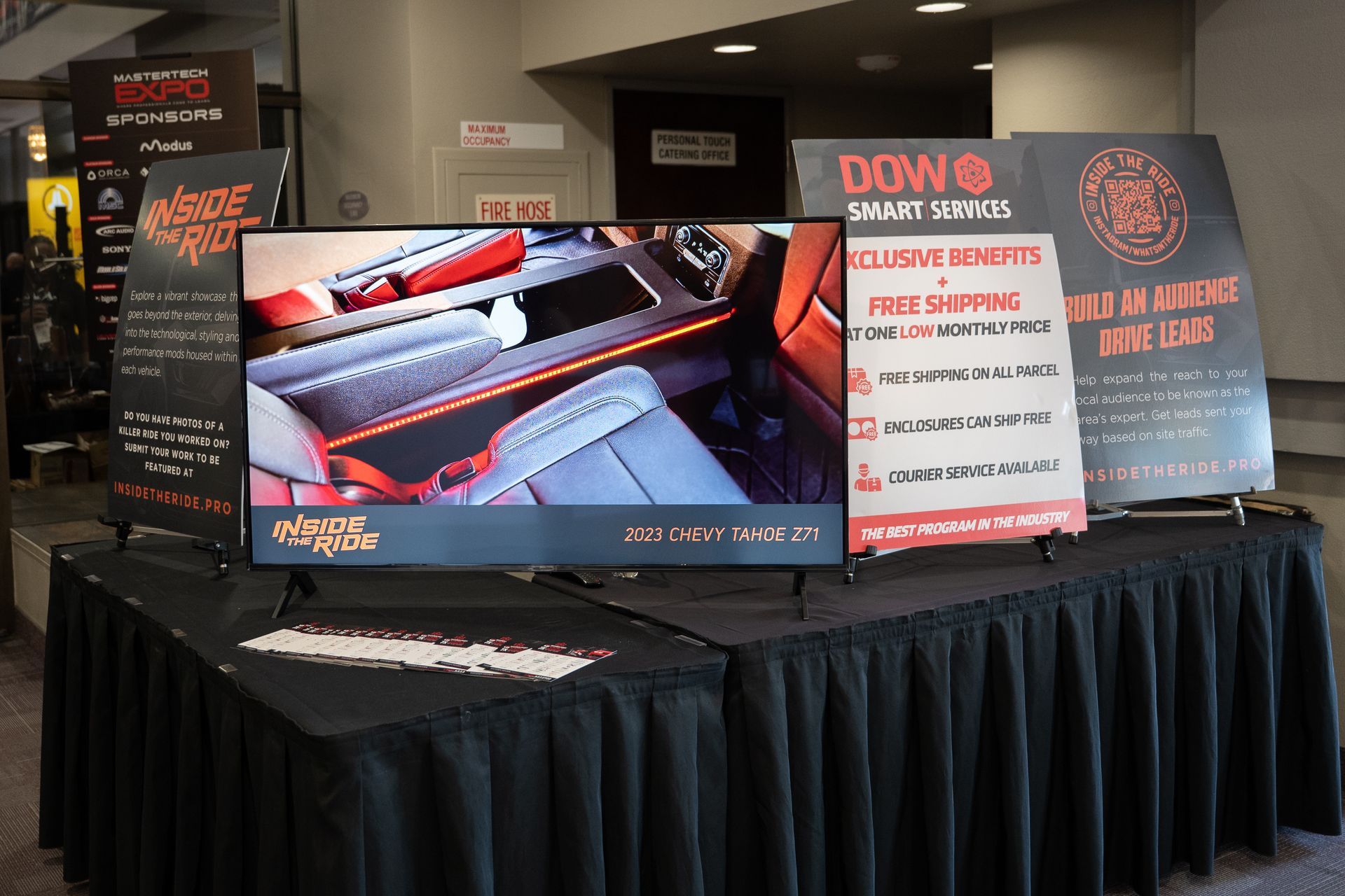 Display table with advertising boards and a TV showcasing a car interior, possibly at a trade show.