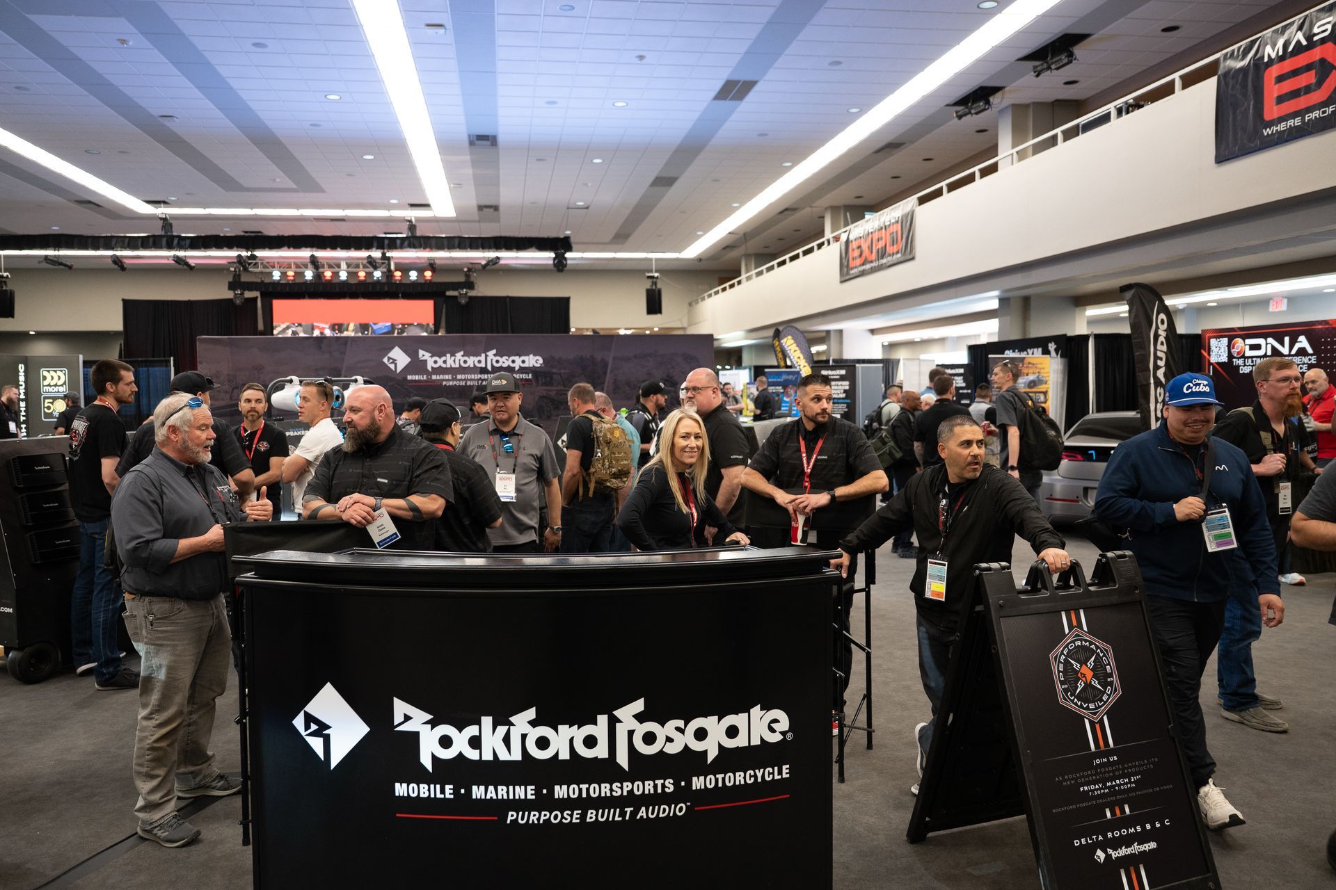 A trade show booth with the Rockford Fosgate logo. People stand around, some wearing lanyards, interacting.