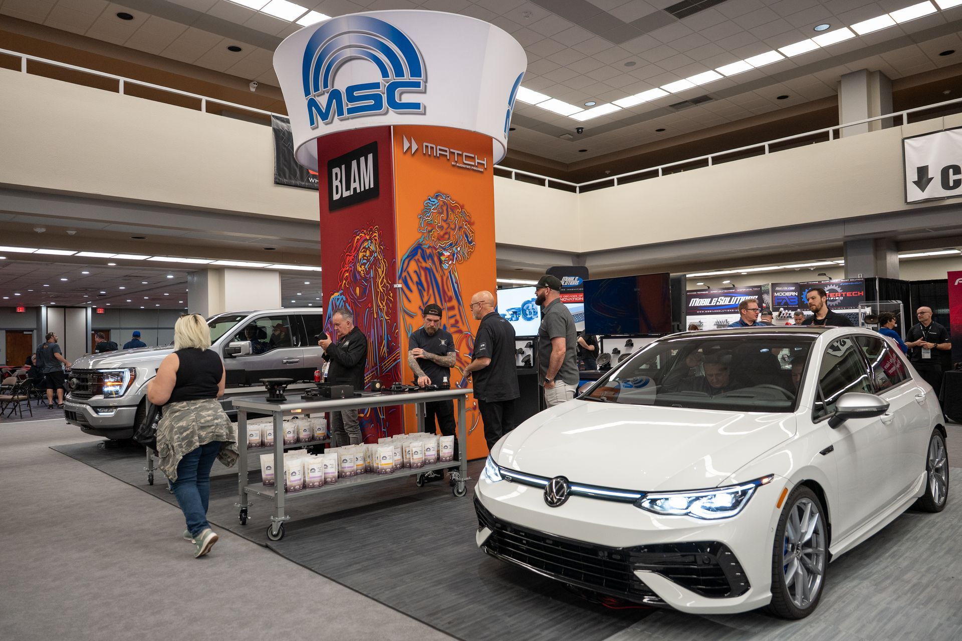 A white Volkswagen car on display at an event. A booth with people, a truck, and a large MSC sign are also visible.