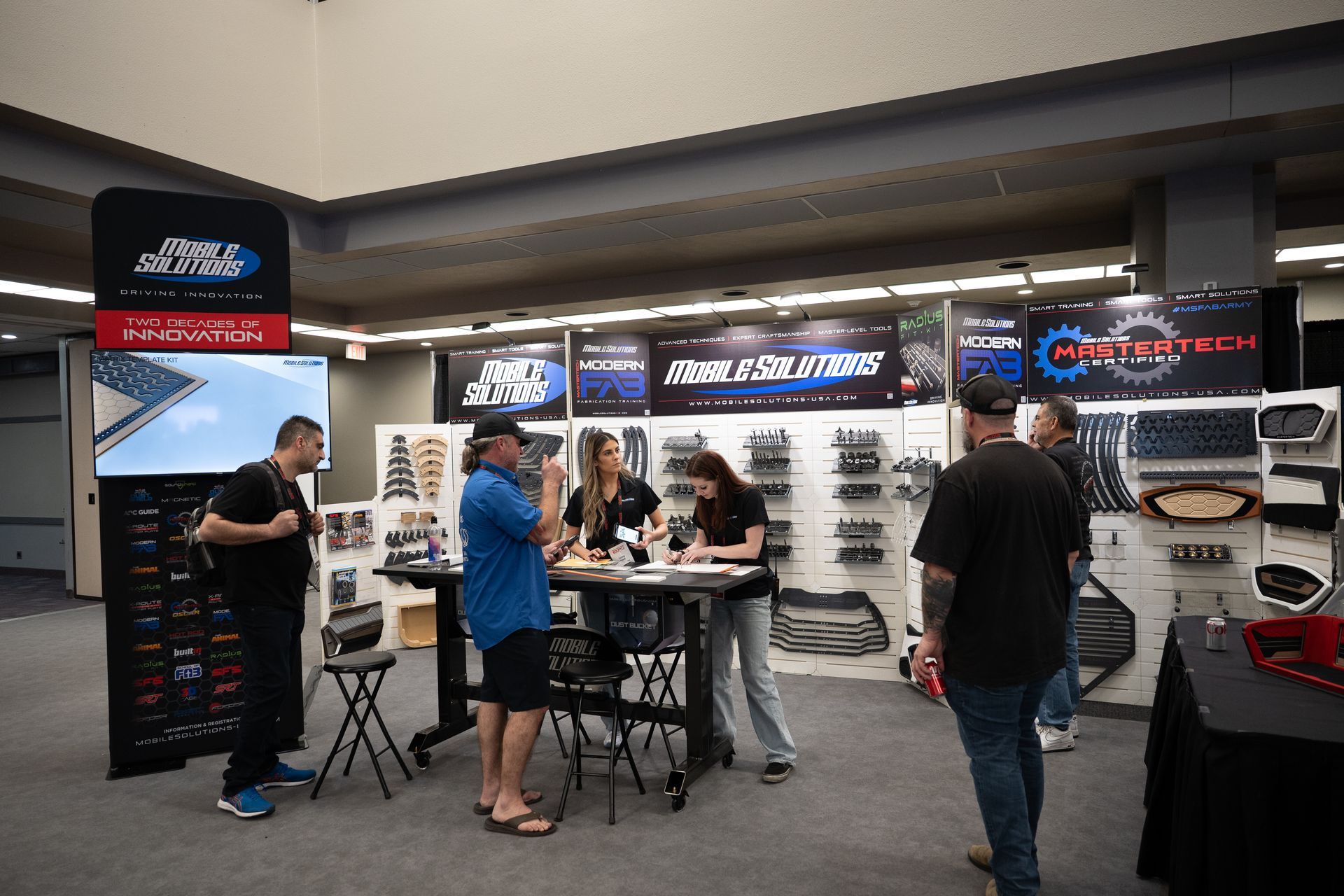 People at a trade show booth with car parts. Products displayed on walls, people at table interacting.
