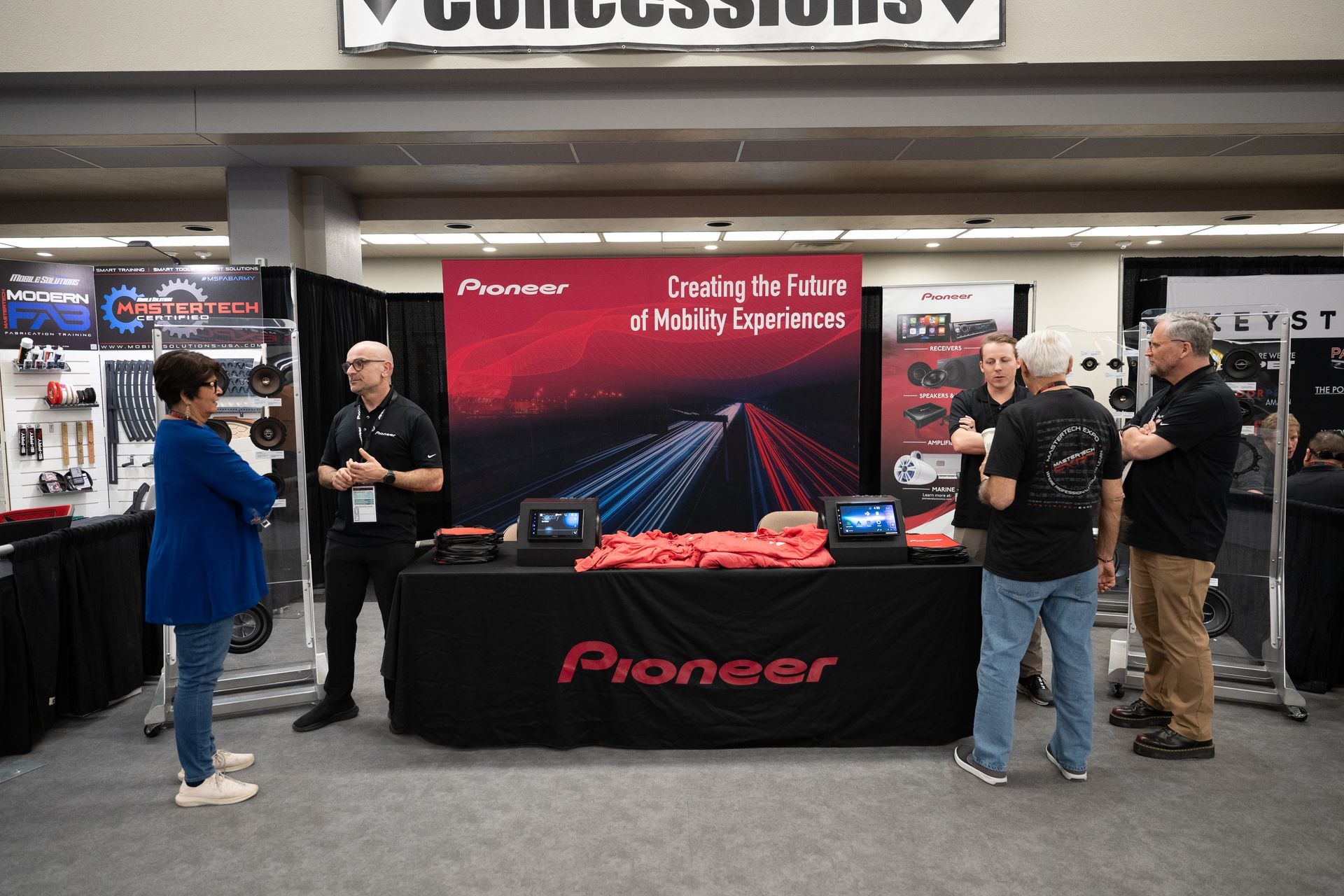 Pioneer booth at a convention: table with products, people talking, red banner, and a concessions sign above.