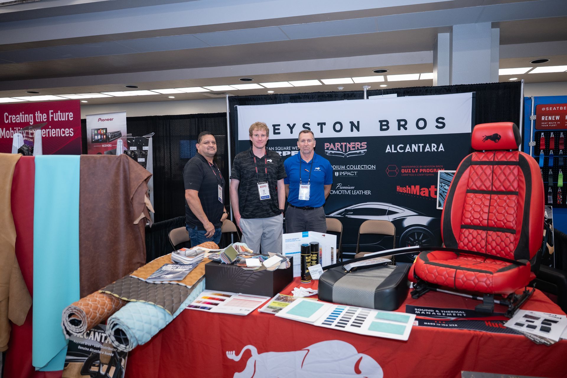 Three men stand at a trade show booth for Keystone Bros, displaying custom car seat with red/black stitching.