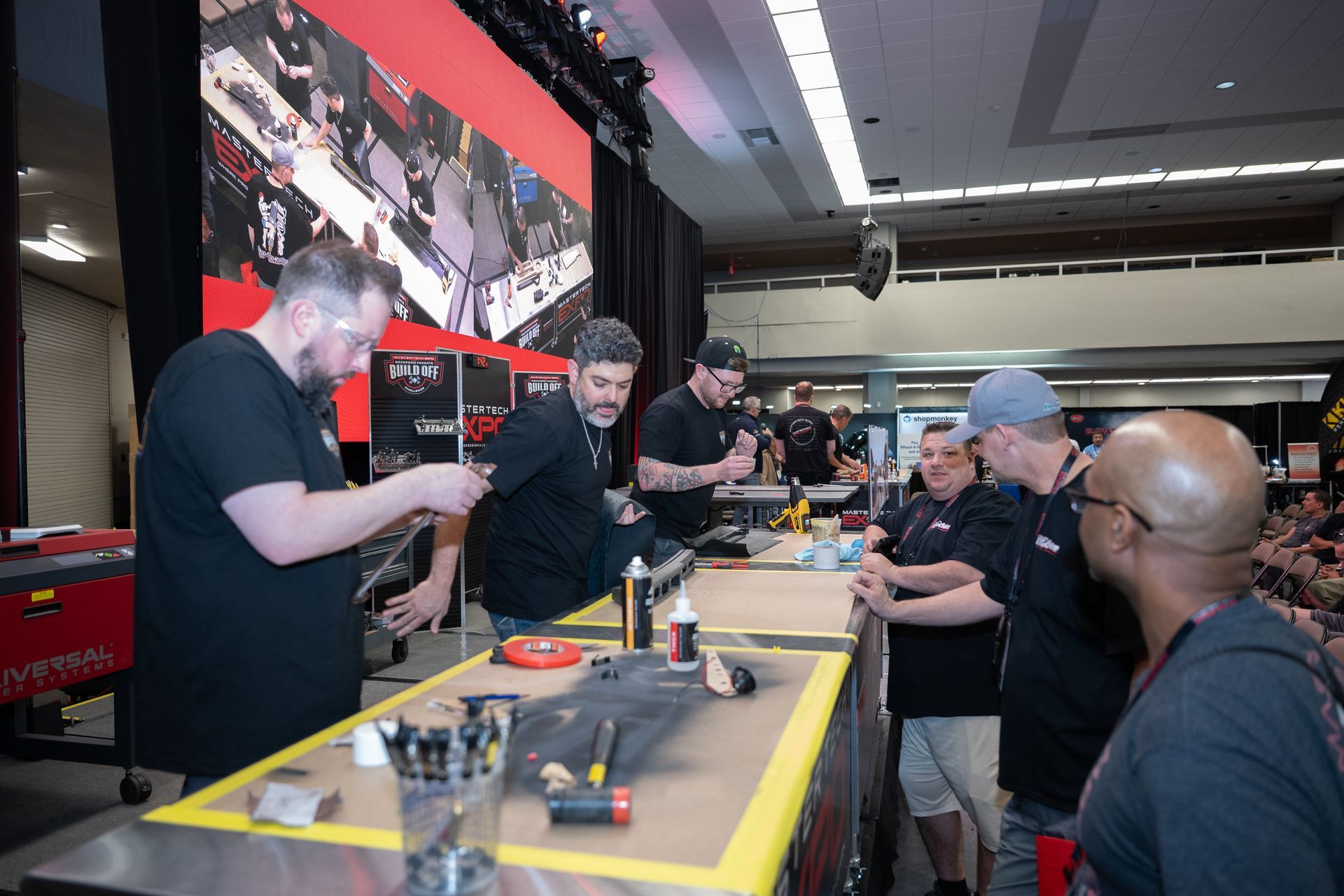 Men at a demonstration, working on objects at a table. A crowd watches. Red and black color scheme.