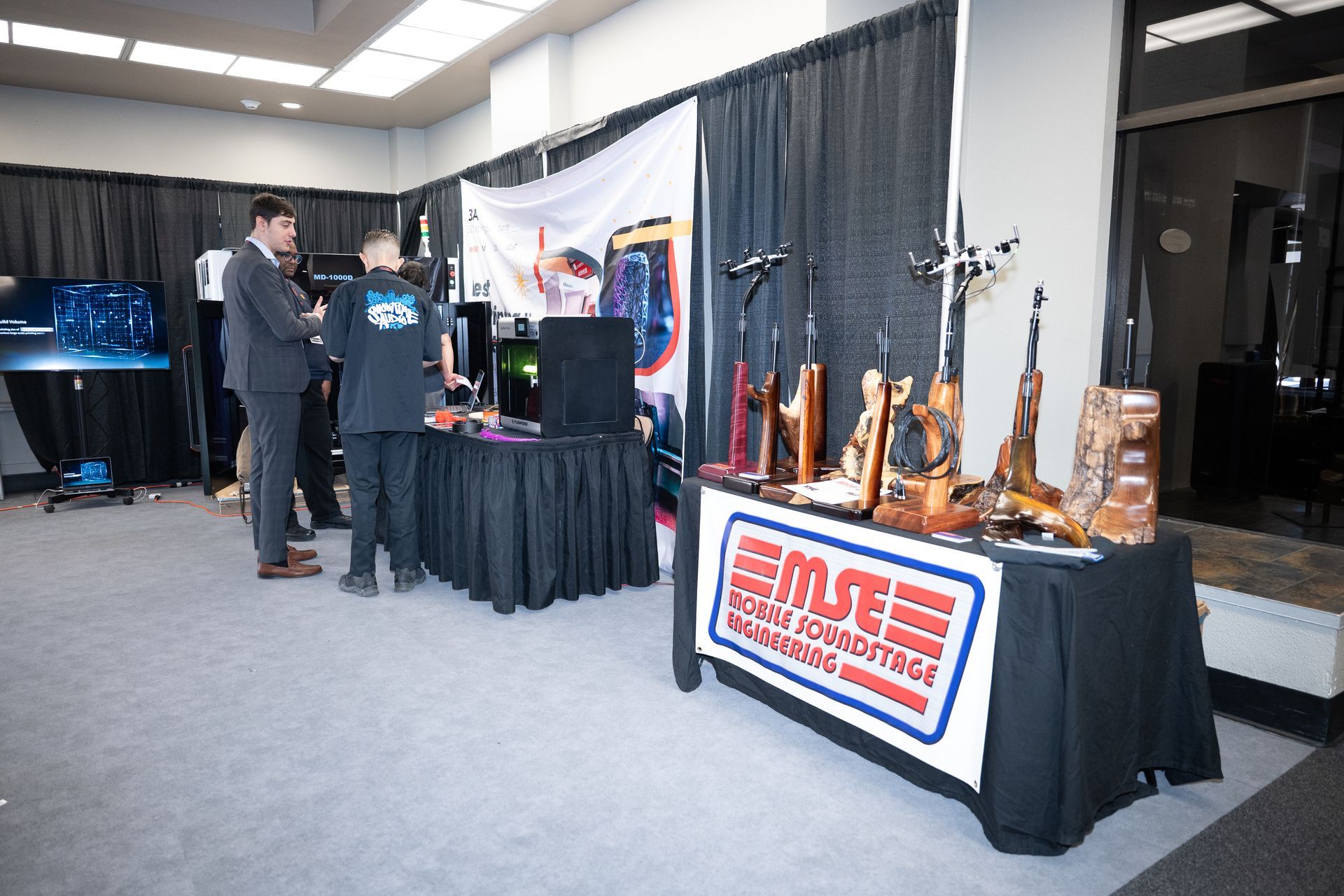 Trade show booth with merchandise on display, two men speaking, and black tablecloths with logos.
