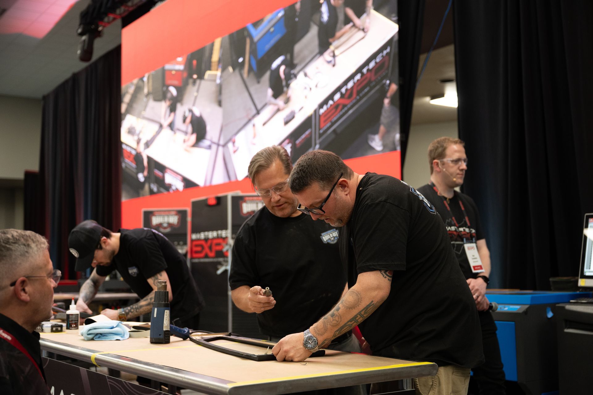 Men assembling a frame at a trade show booth, with a large screen displaying an overhead view.
