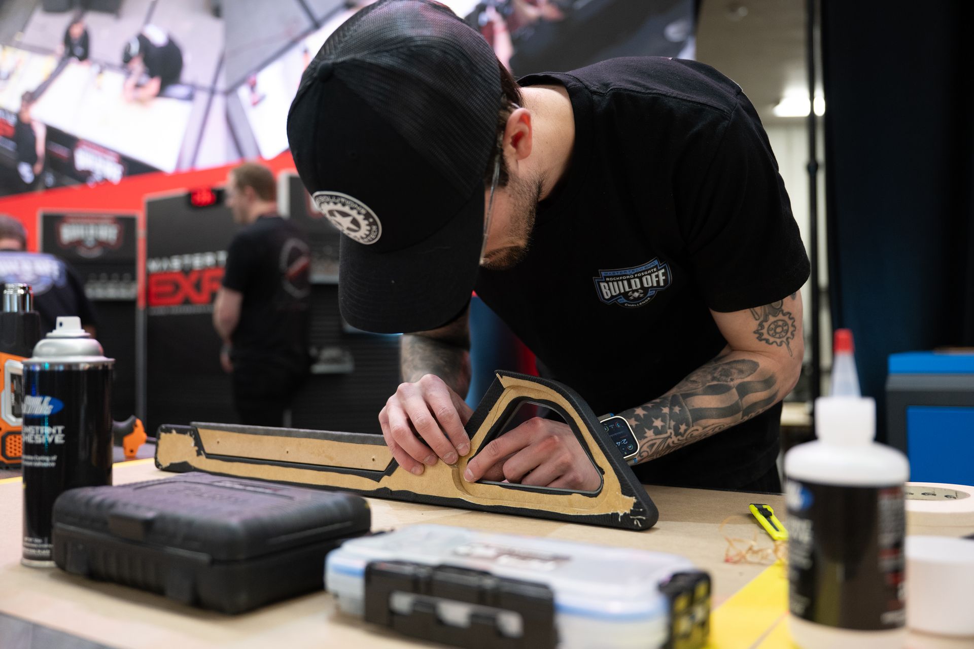 A person wearing a black cap and shirt working on a project, possibly assembly, at a table.