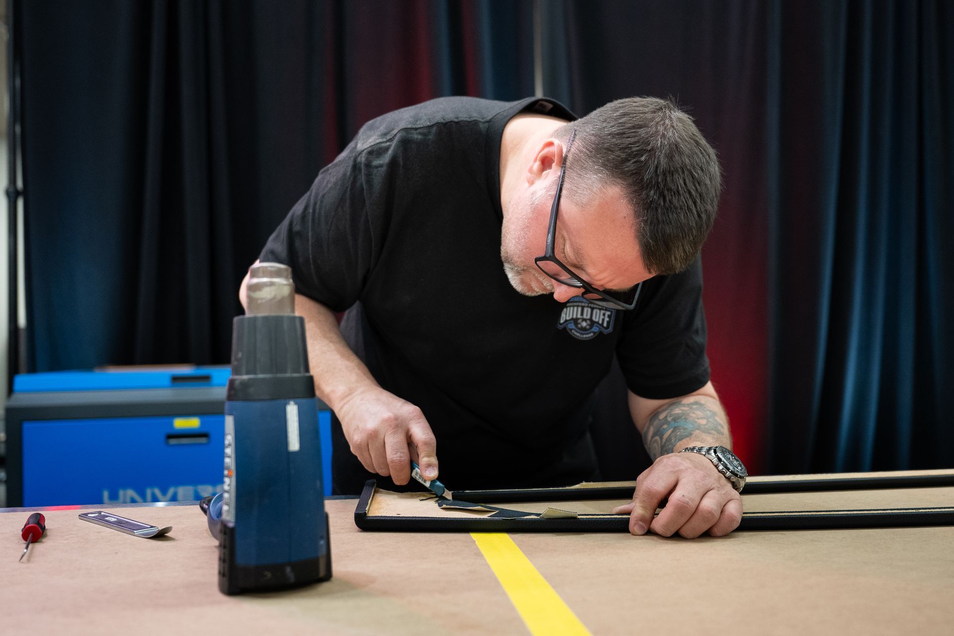 Man with magnifying glasses and tools, working on a black frame. Dark shirt, tattoos, indoor setting.