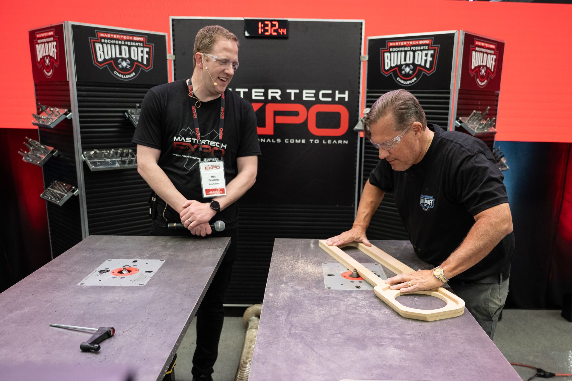 Two men at a woodworking expo. One uses a router on a wooden frame. The other watches, holding a microphone.