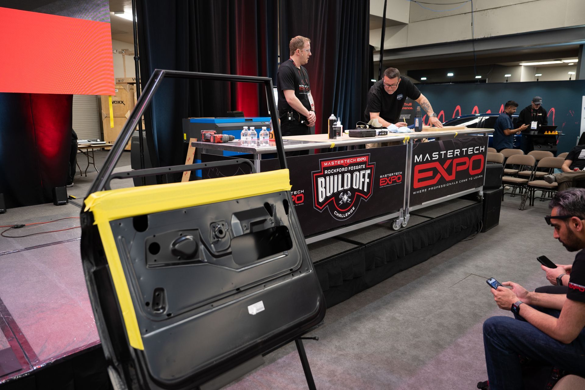 Workshop with a car door on display, two men at a table with tools and expo banners.