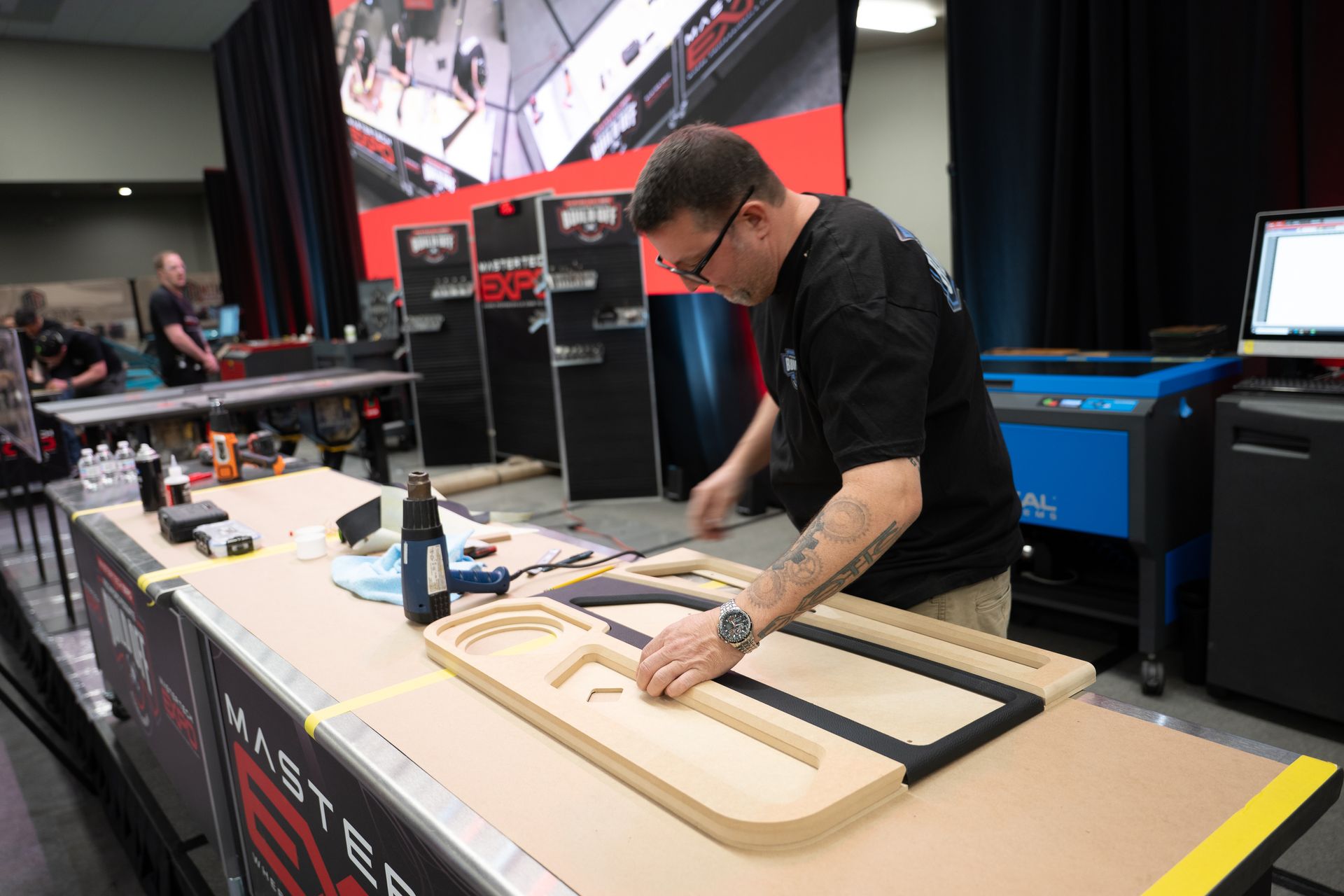 Man working on a car door panel at a trade show; tools and equipment on the table.