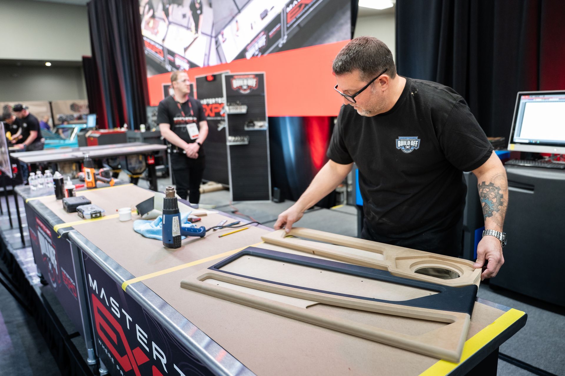 Man working on a car door panel at a trade show; tools and a computer on a table.