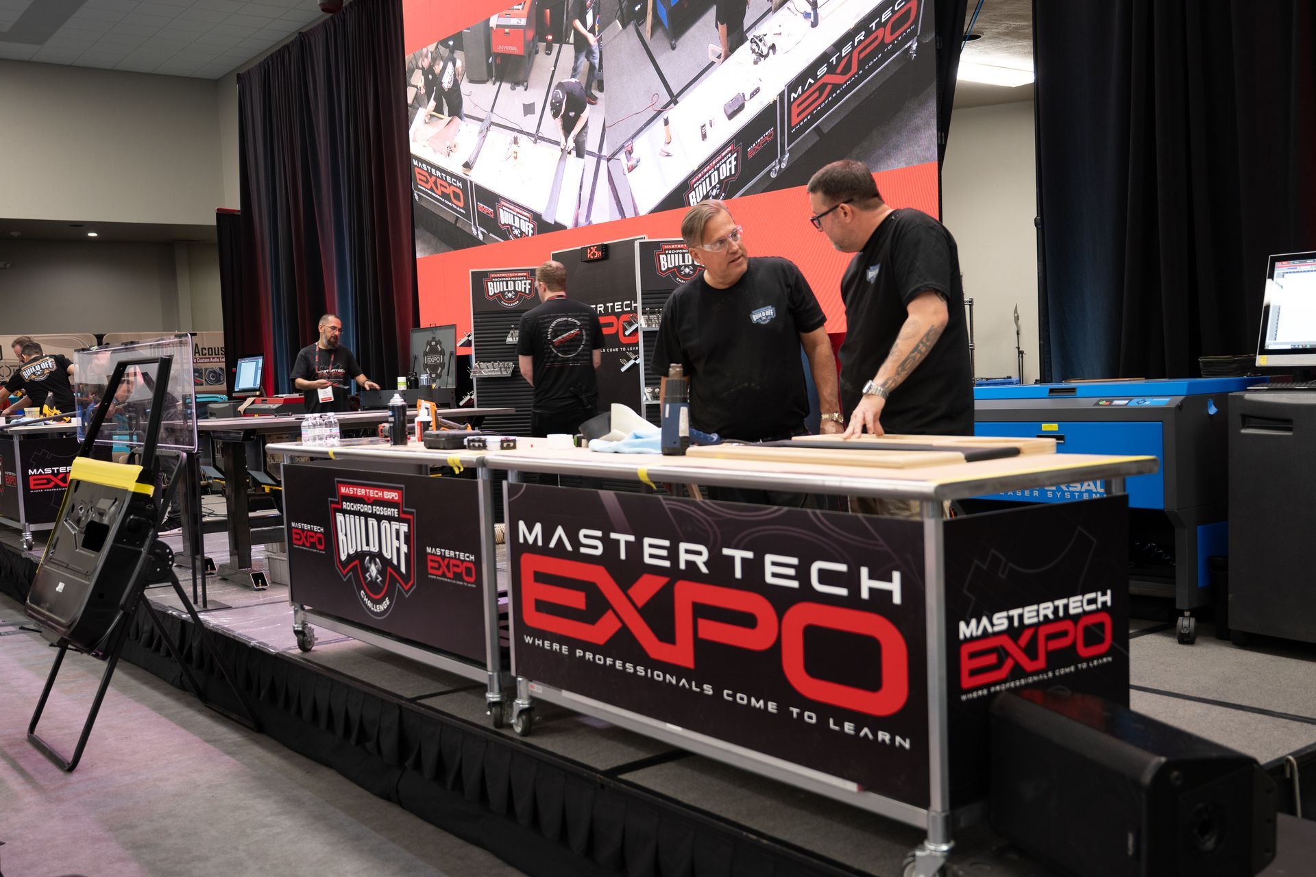 People interacting at the MasterTech Expo. Men at a table with tools; a large screen is in the background.