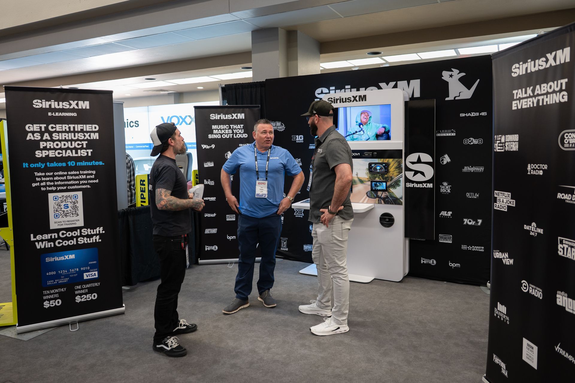 Three men converse at a SiriusXM booth at a trade show; black and white display panels are behind them.