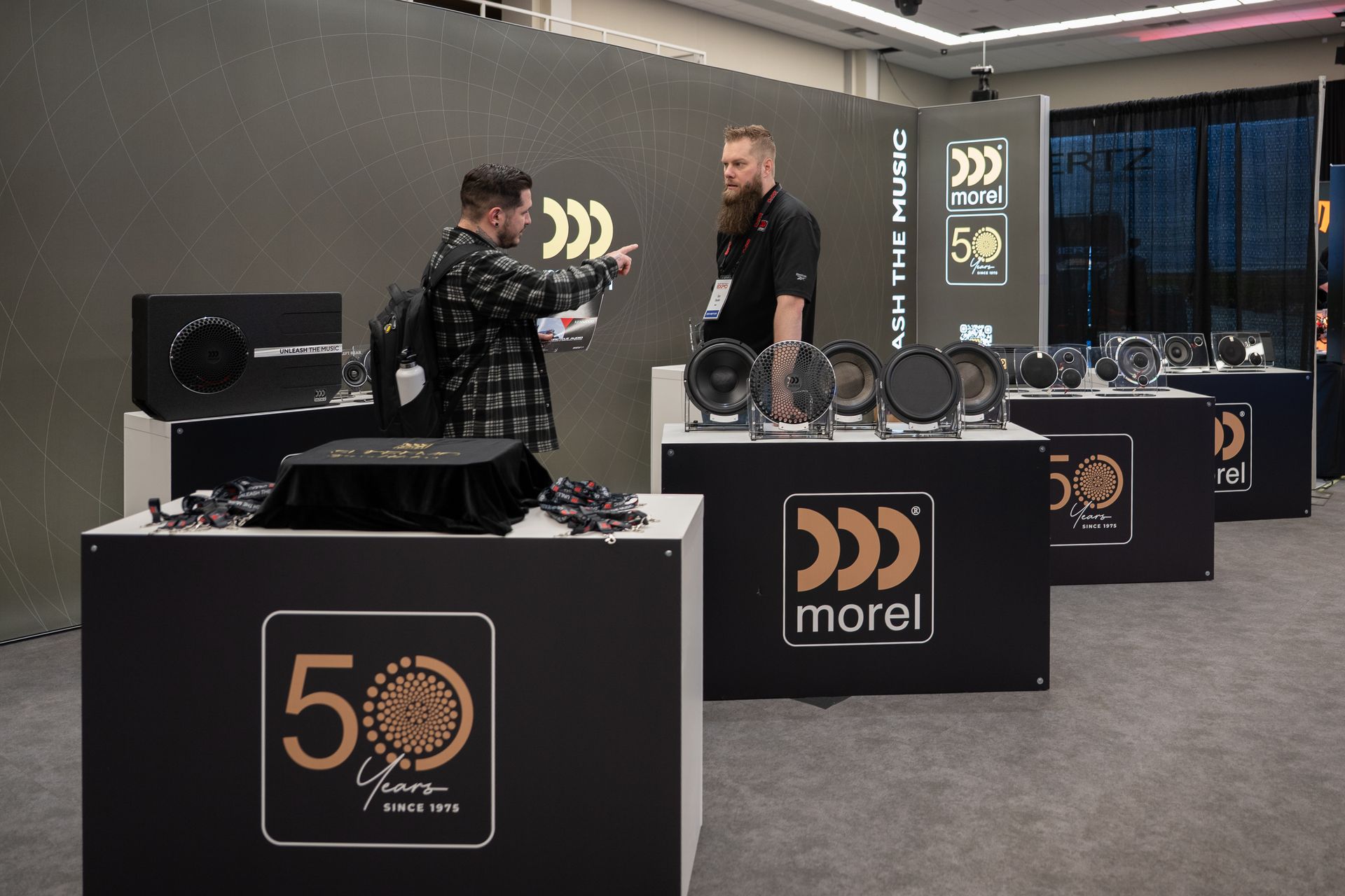 Two men at a Morel audio products display. One gestures, the other listens. Products are visible on tables.