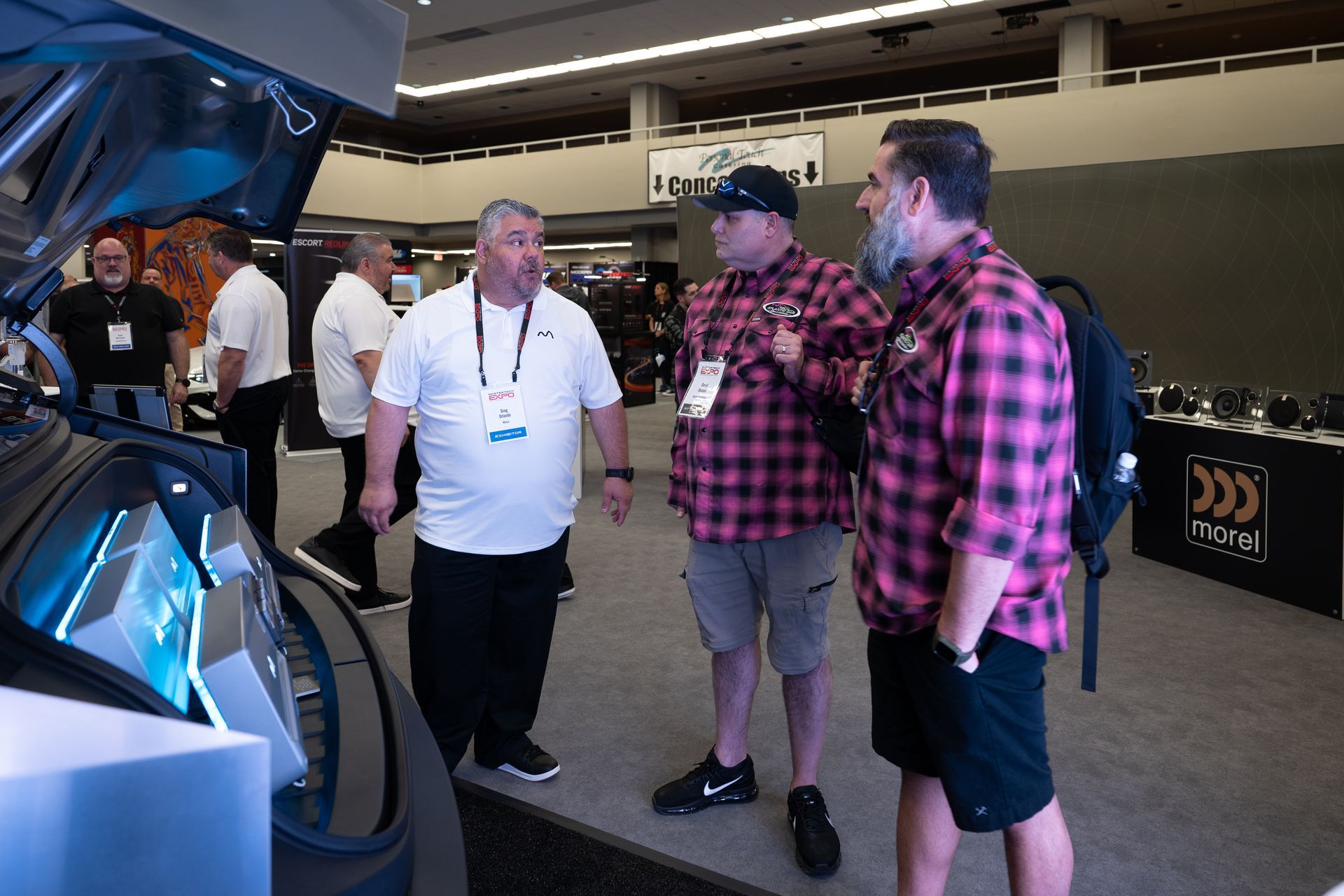 Three men at a car show, inspecting a vehicle. One in a plaid shirt with a beard, another in a cap and the other in a white shirt.