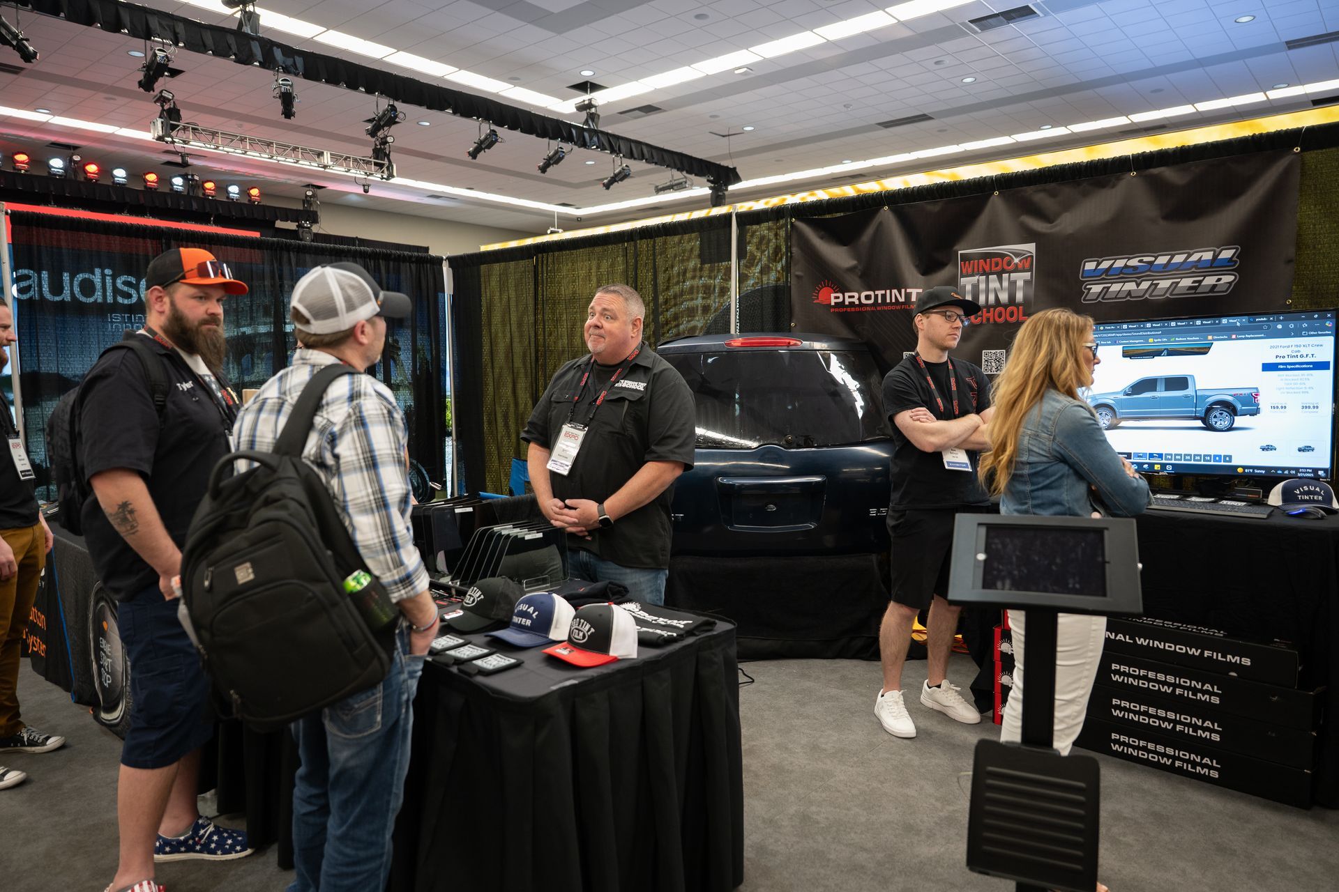 People at a trade show booth with a black truck, display, and merchandise.