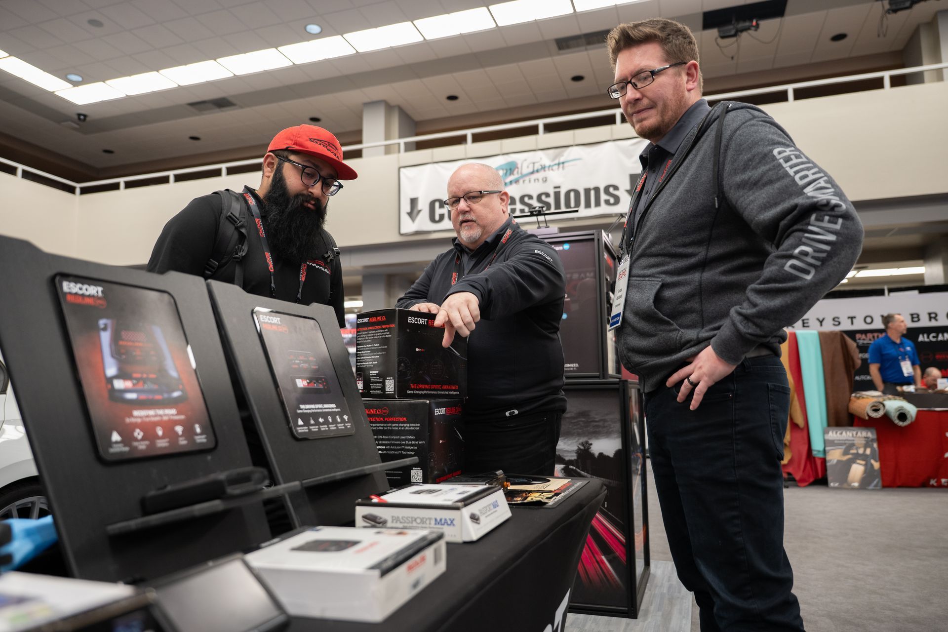 Three men at a trade show booth, looking at display screens. One points. Boxes of products are on a table.