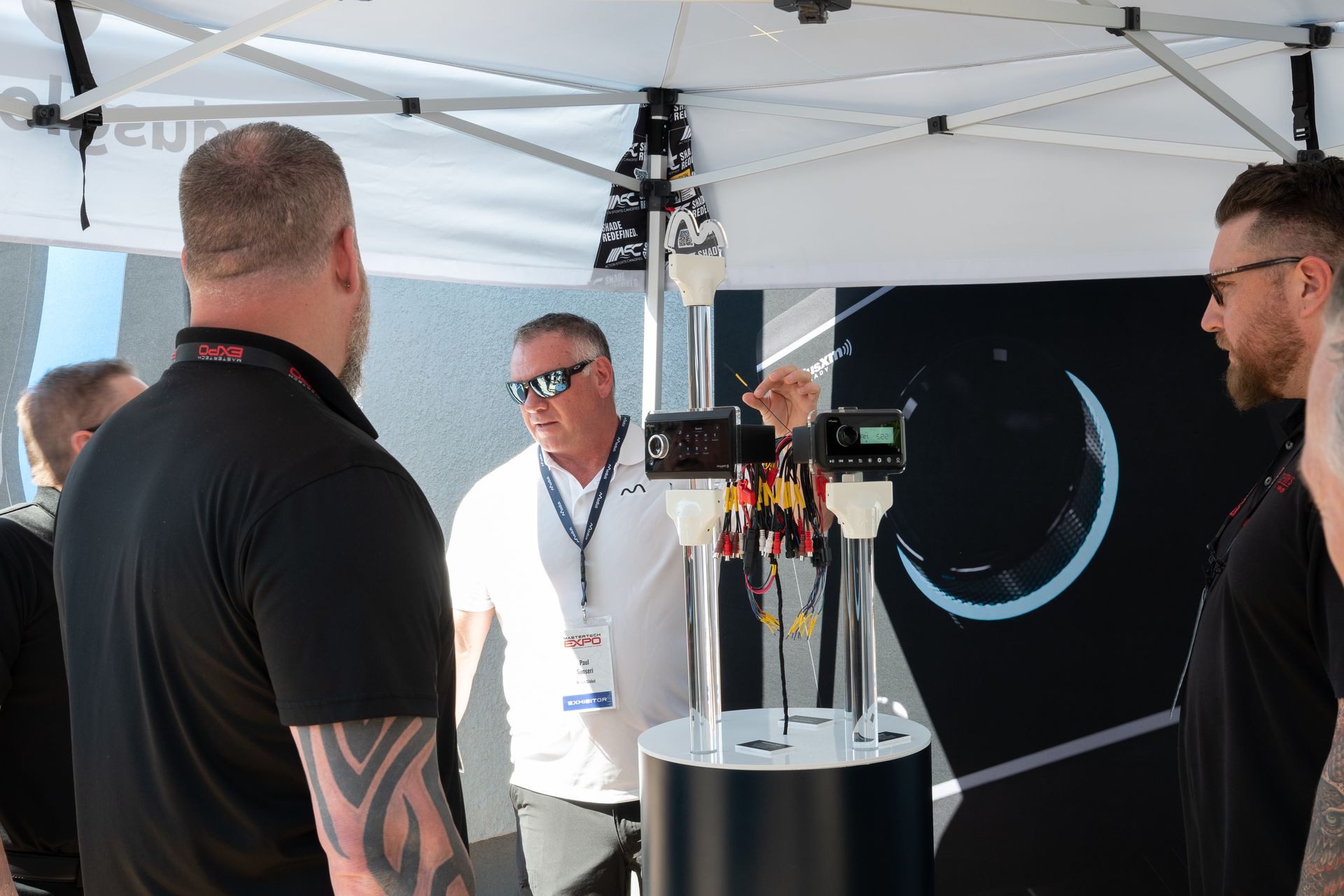 People examining electronic devices at a display booth under a tent; one person points to them.
