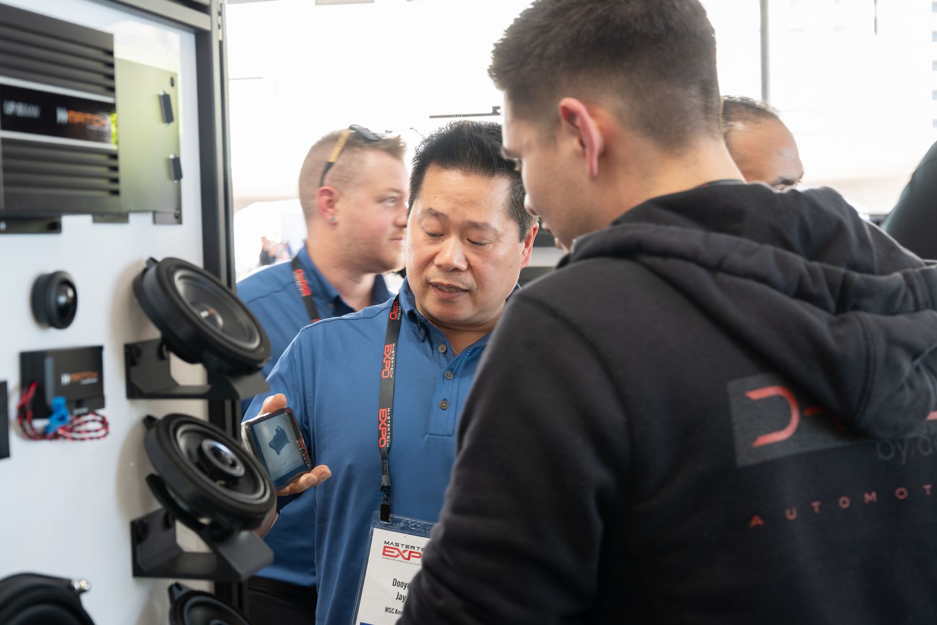 Man showing car speaker to another at a trade show booth, other men in background.