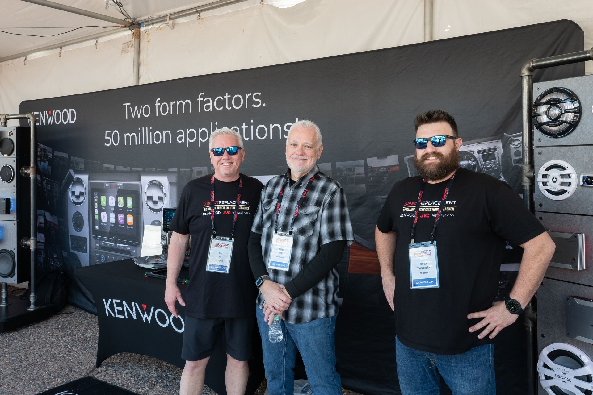 Three men stand in front of a Kenwood display. The backdrop has product images and text. The men wear sunglasses and name badges.