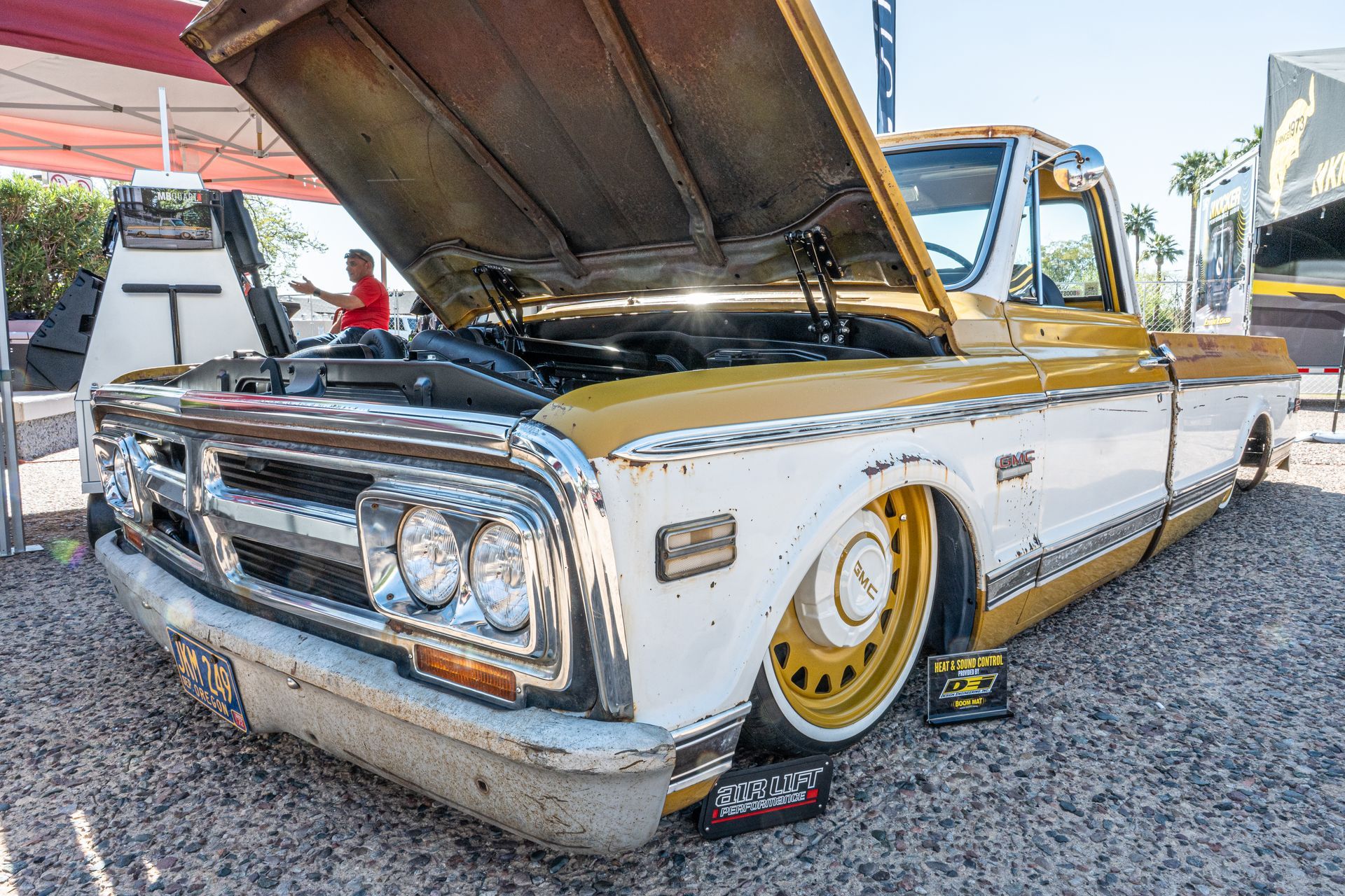 Yellow and white classic truck with its hood up, parked at an outdoor car show.