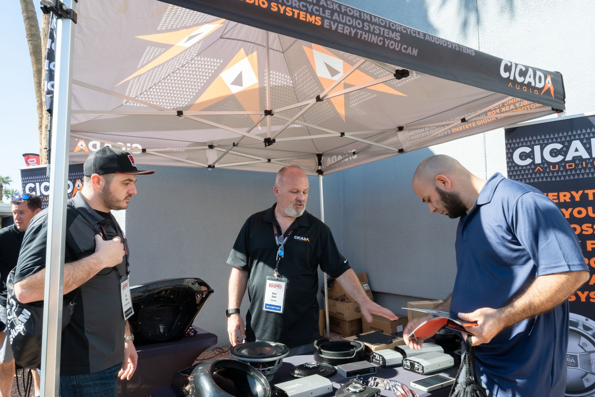 Three men at a Cicada booth, examining motorcycle audio systems. Outdoor setting, tent overhead.
