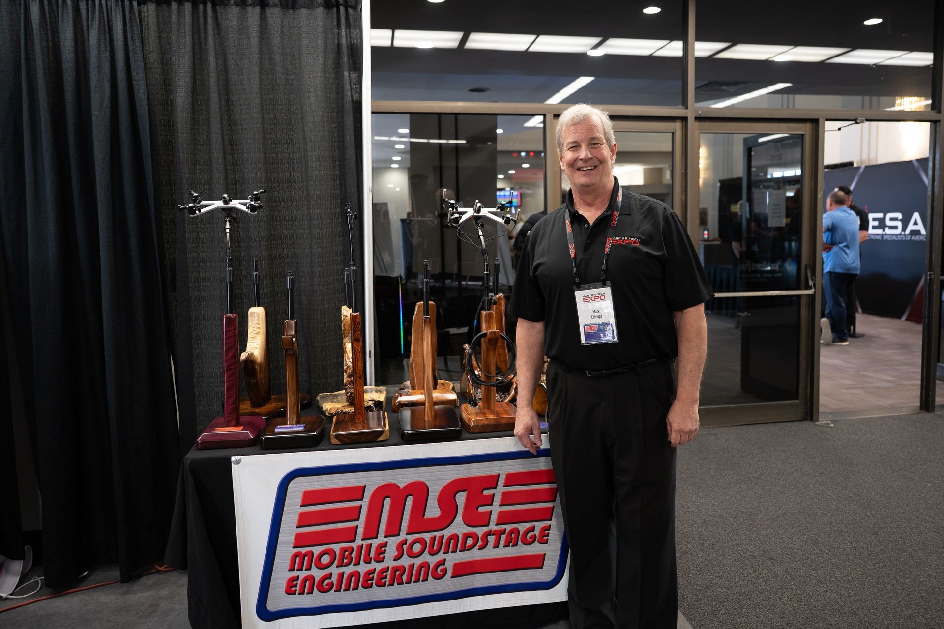 Man smiling next to a display of wooden musical instrument components at a trade show booth, 