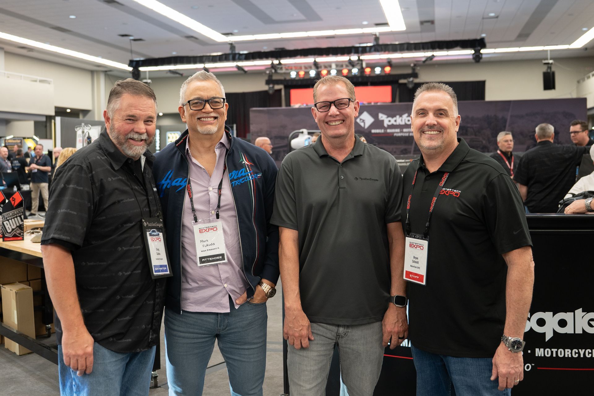 Four men smiling at a trade show booth, indoors with equipment visible behind them.