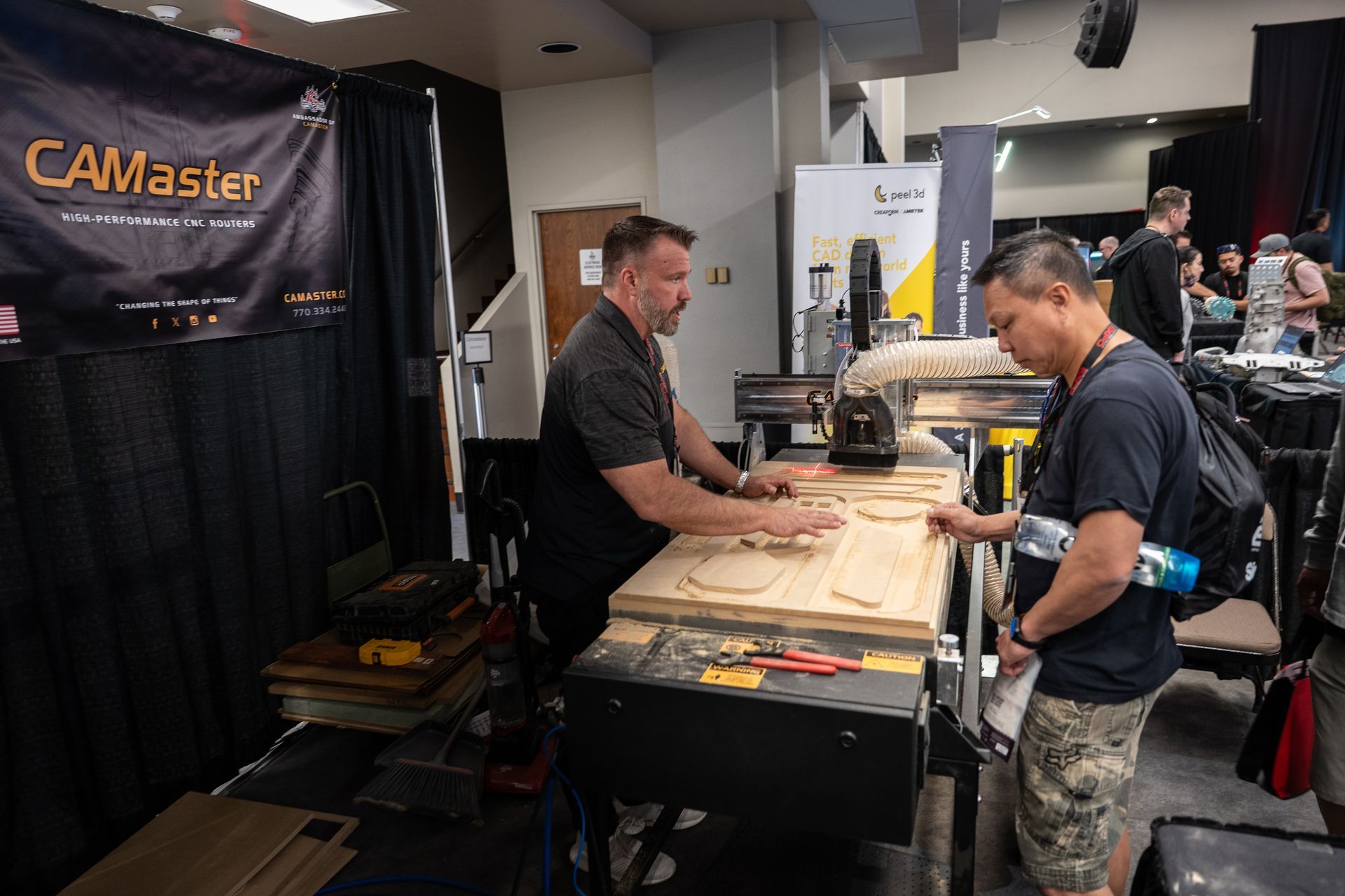 Man demonstrating a CNC router to another man at a trade show booth, CAMaster banner in the background.