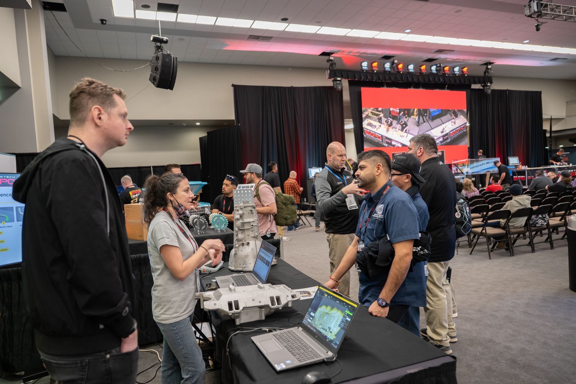 People gather at a tech event. One woman explains a device at a table. Laptops, and a stage are in the background.