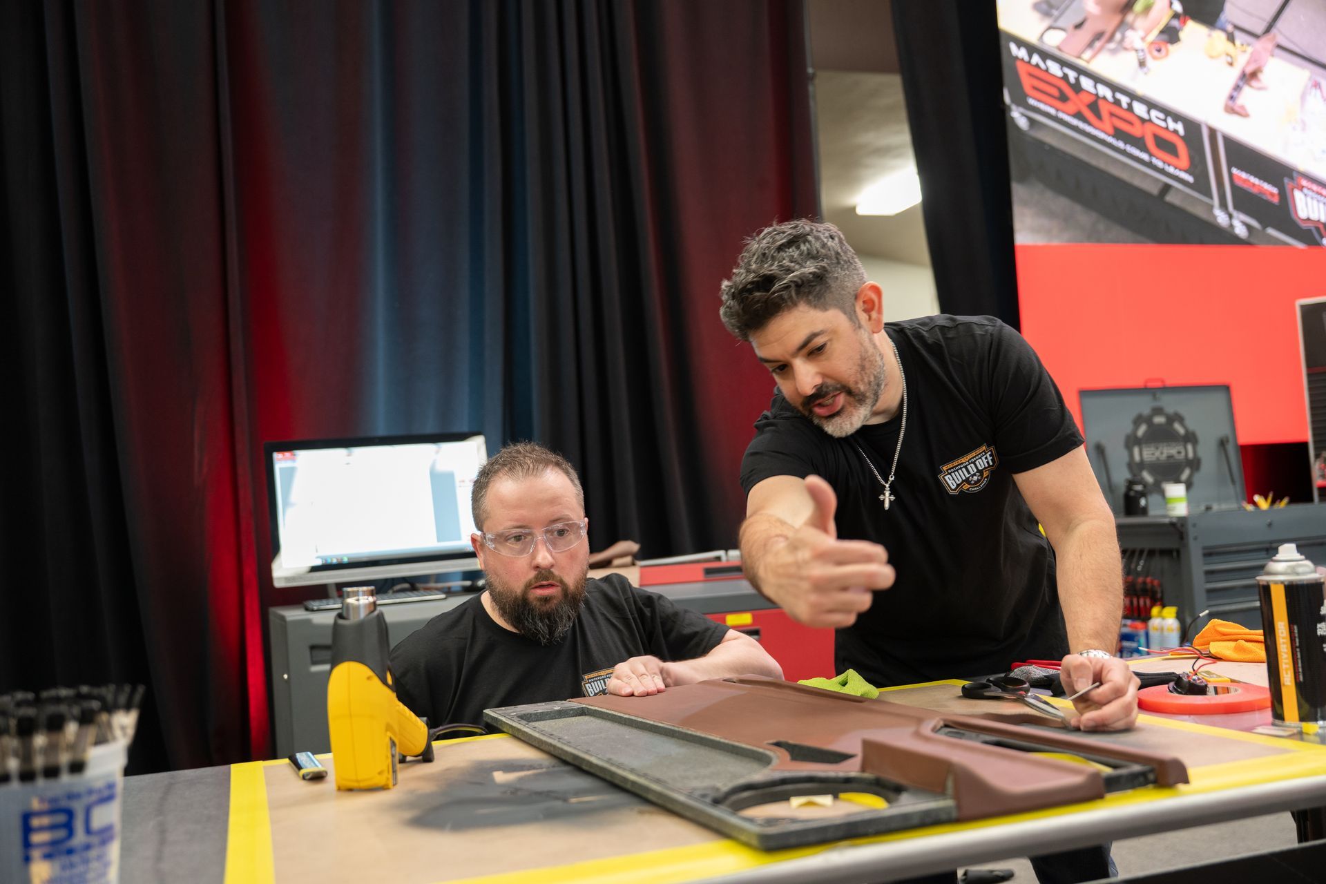 Two men working on a car door panel, one pointing, the other looking on, in a workshop setting.