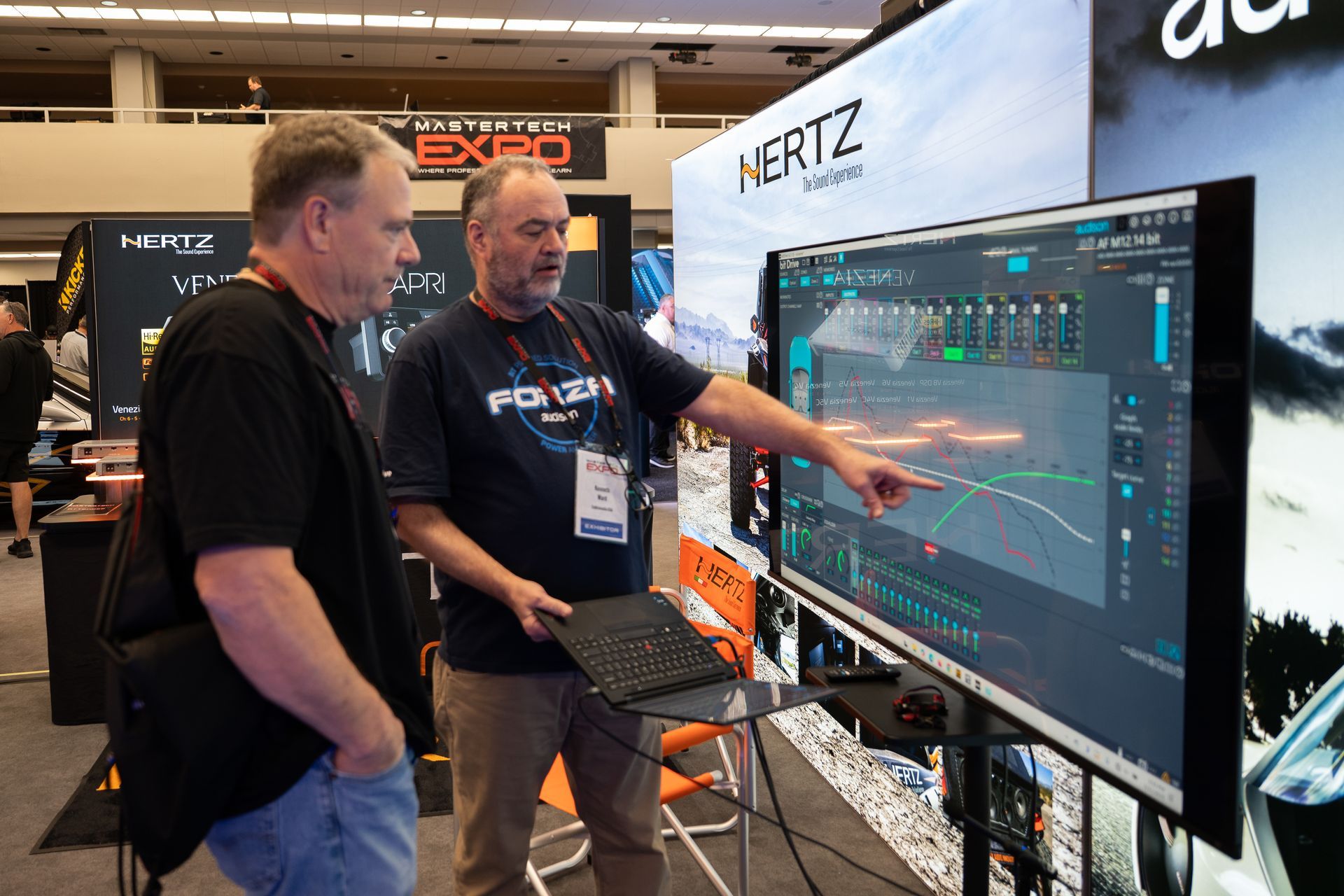 Two men looking at a large screen with sound wave graphs. One points at the screen, a trade show setting.