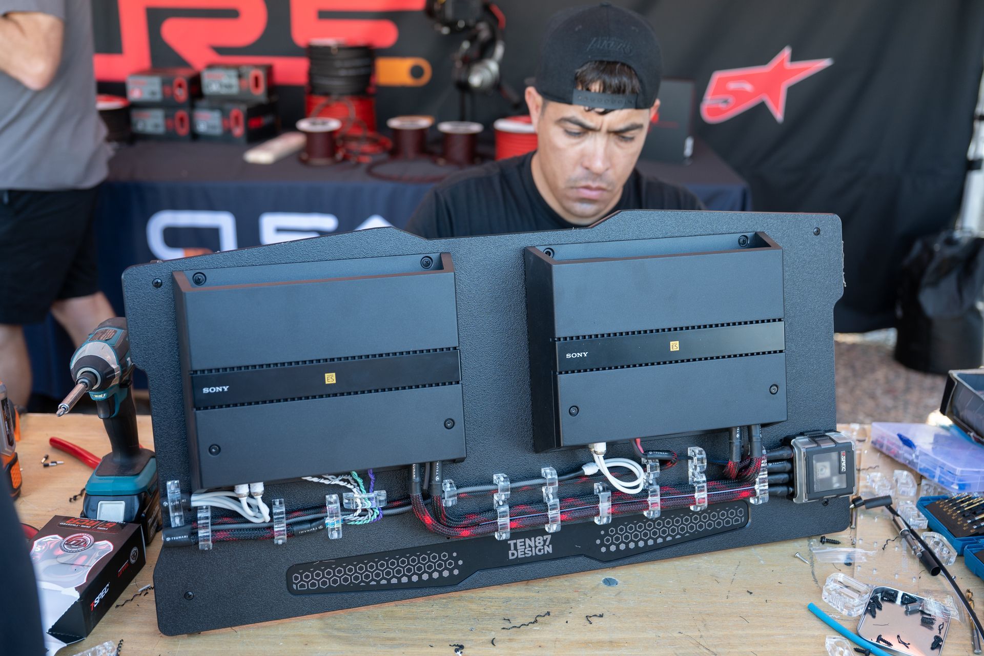 Man assembling car audio components with tools on a table outdoors, near display of speakers.