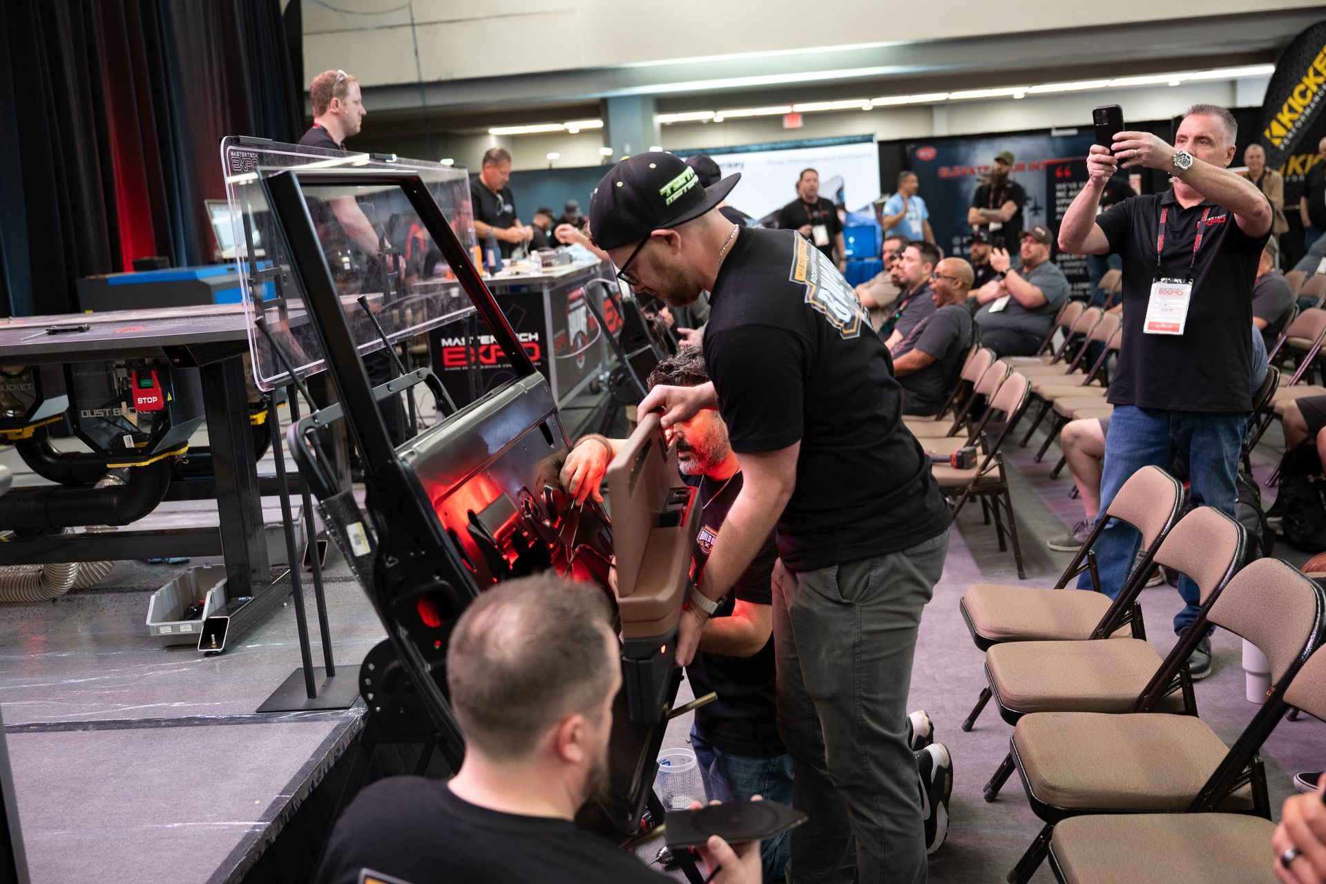 People working on a robot in a competition setting, surrounded by spectators.