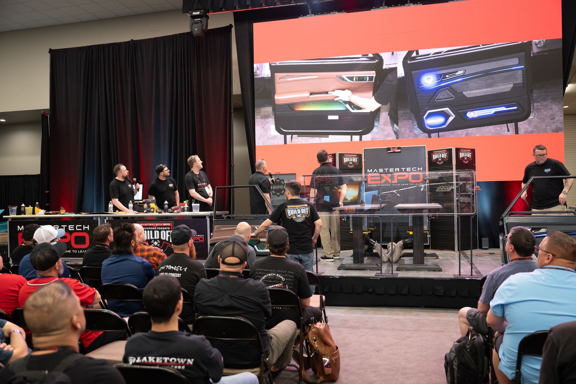 Gamestop Expo: Presenters on stage with computers, display screen; audience watching.