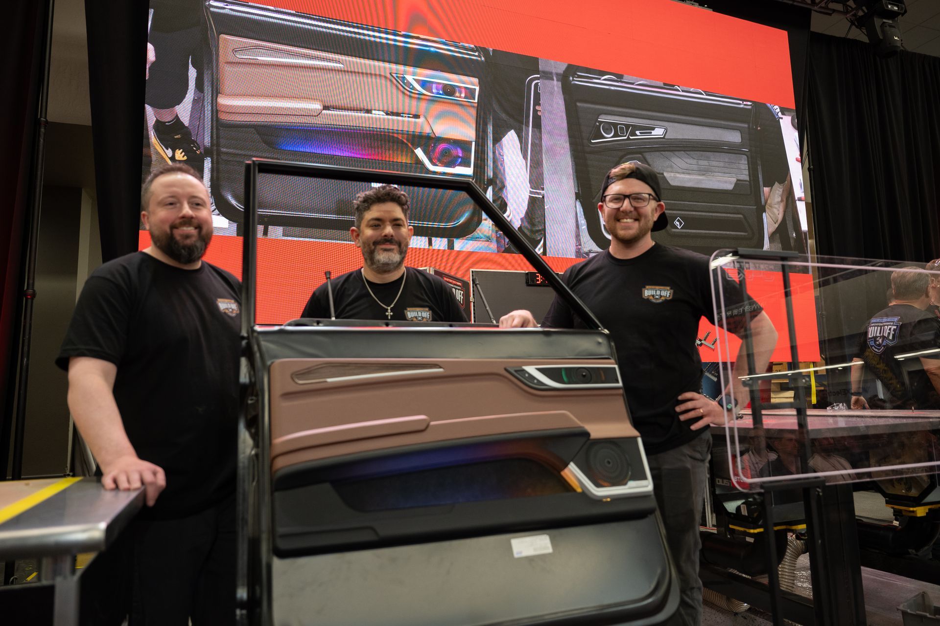 Three men pose with a large car door mockup. Background is a digital display of car interior.