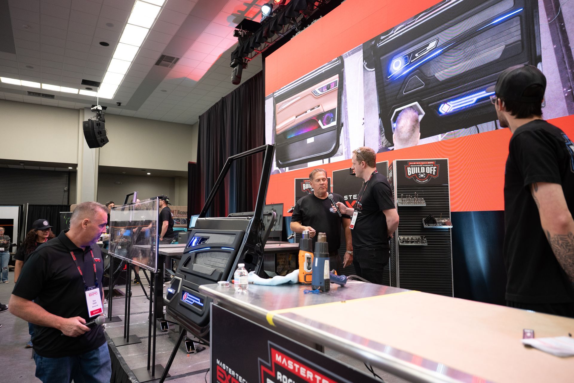 People at a display booth. A large screen shows images. Several people look at displays and converse.