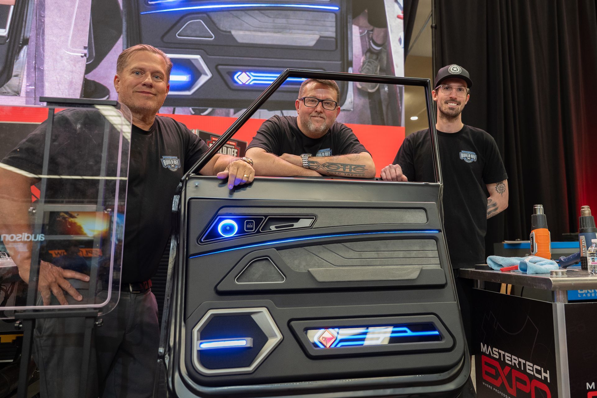 Three men pose with a custom car door at an expo. The door is black with blue lights.