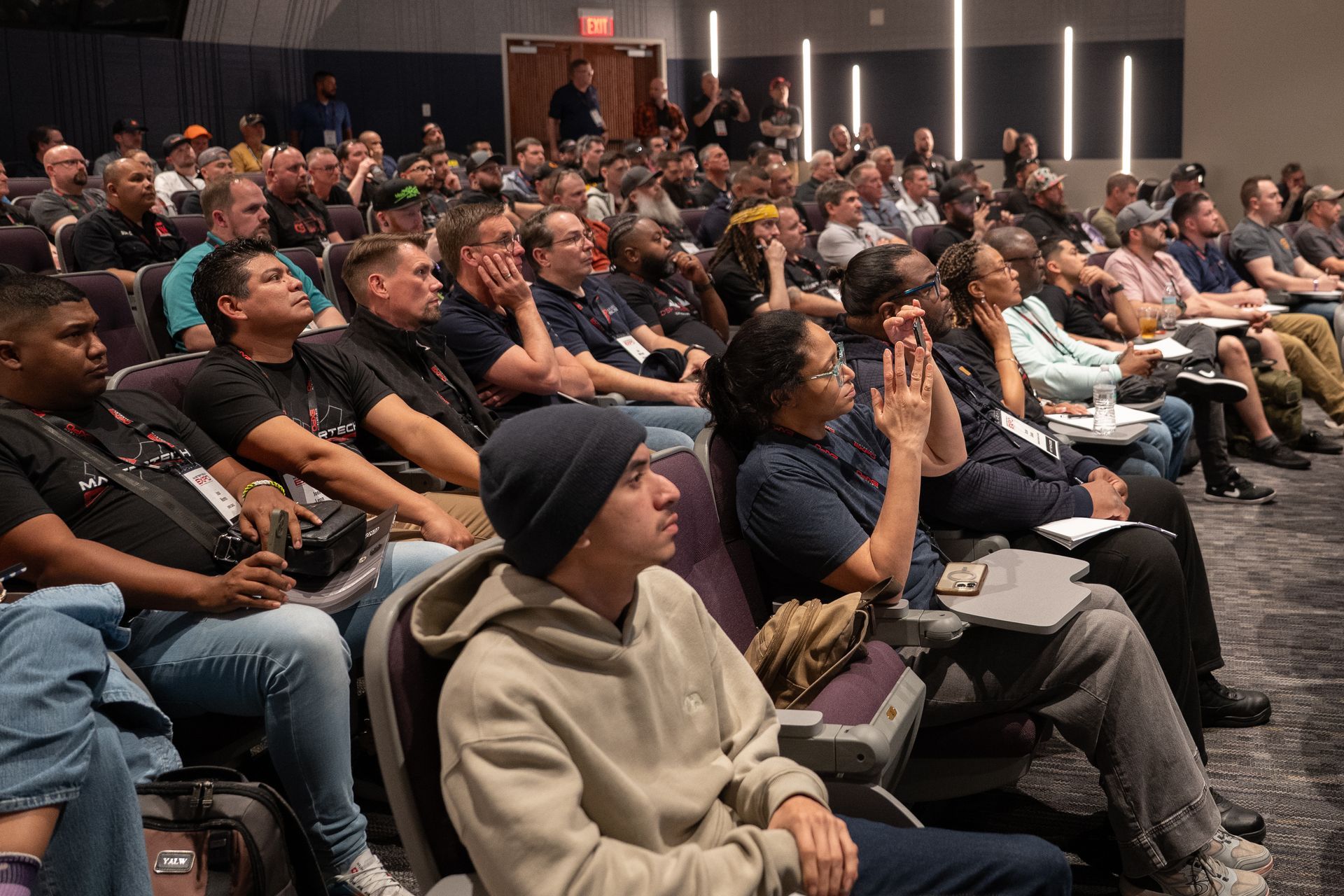 Audience in a conference room; people listen attentively, some take notes.