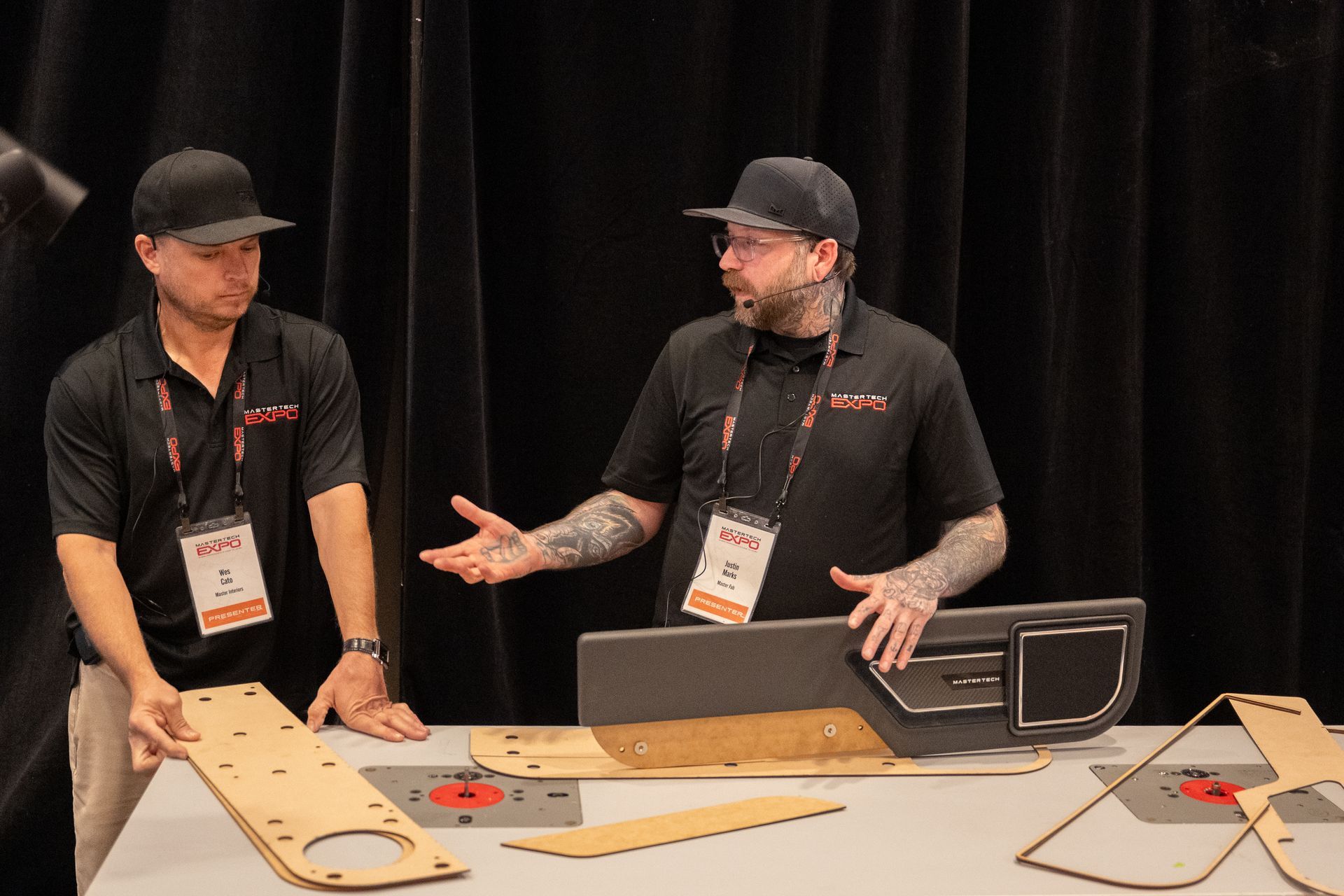 Two men at a booth demonstrate car interior parts. One gestures, other holds a panel. Tools and parts are on table.