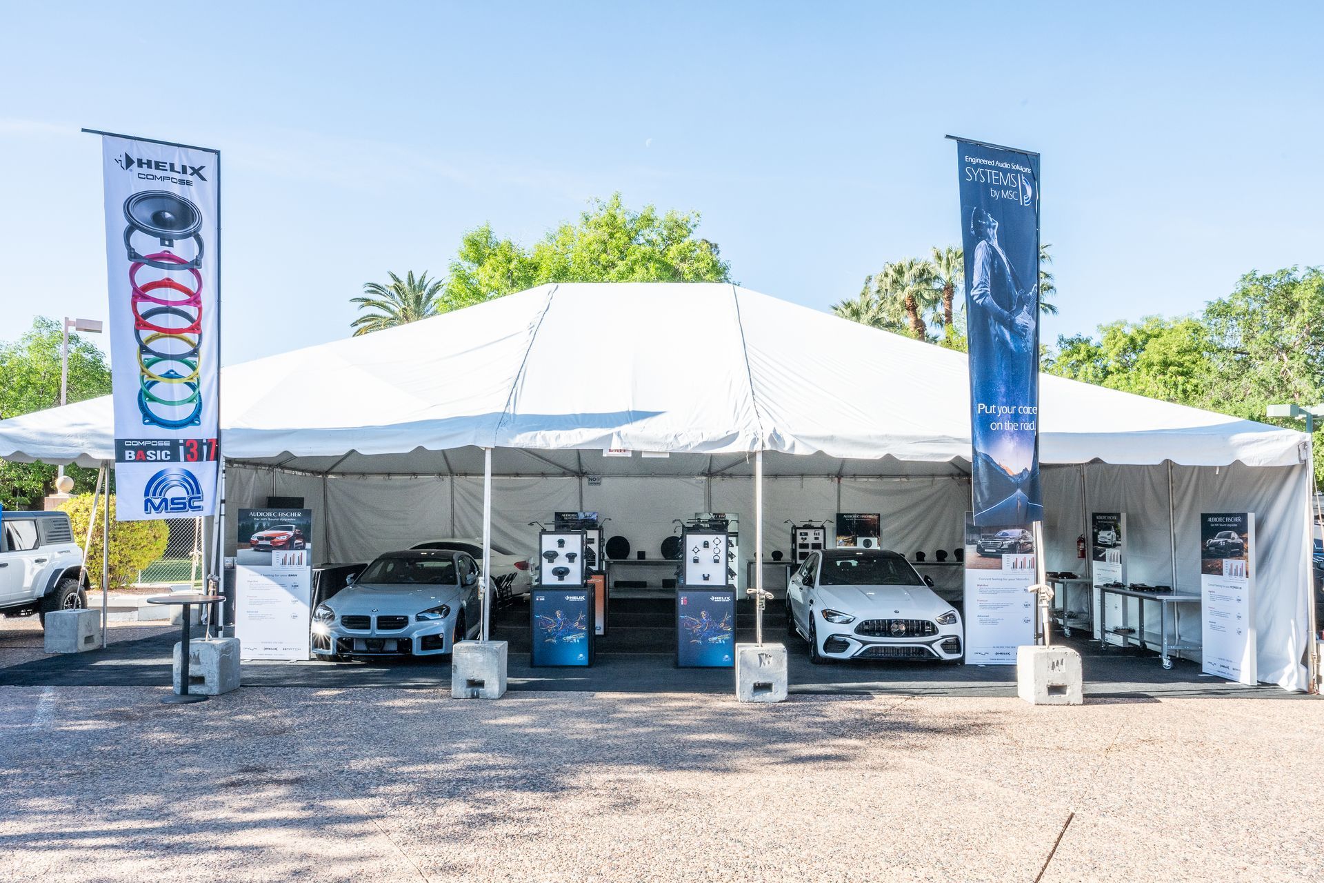White tent with cars on display, sponsored by banners on a sunny day.